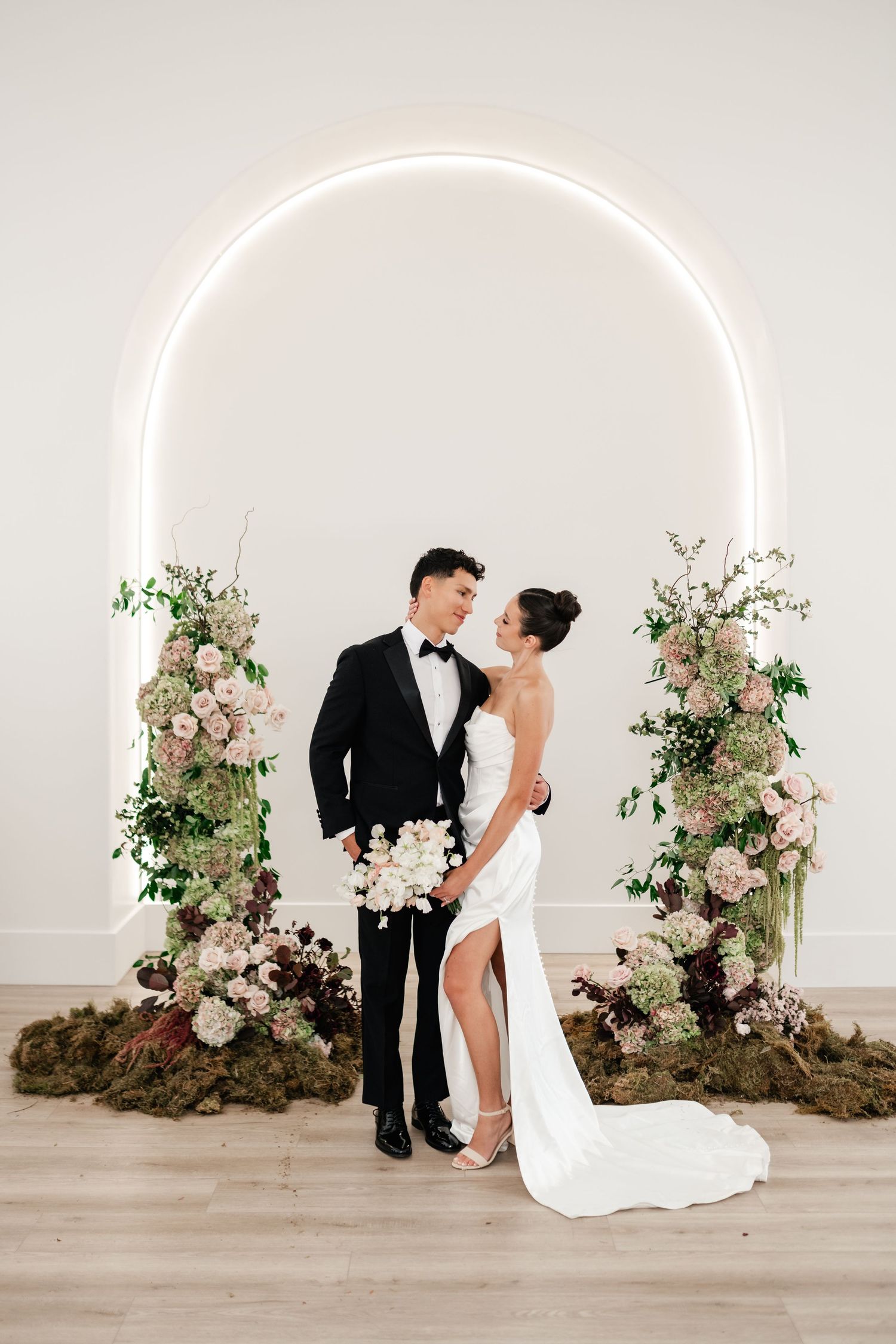 Elegant wedding couple stands under illuminated circular arch with romantic floral arrangements in white and pink. Moments on Main preferred