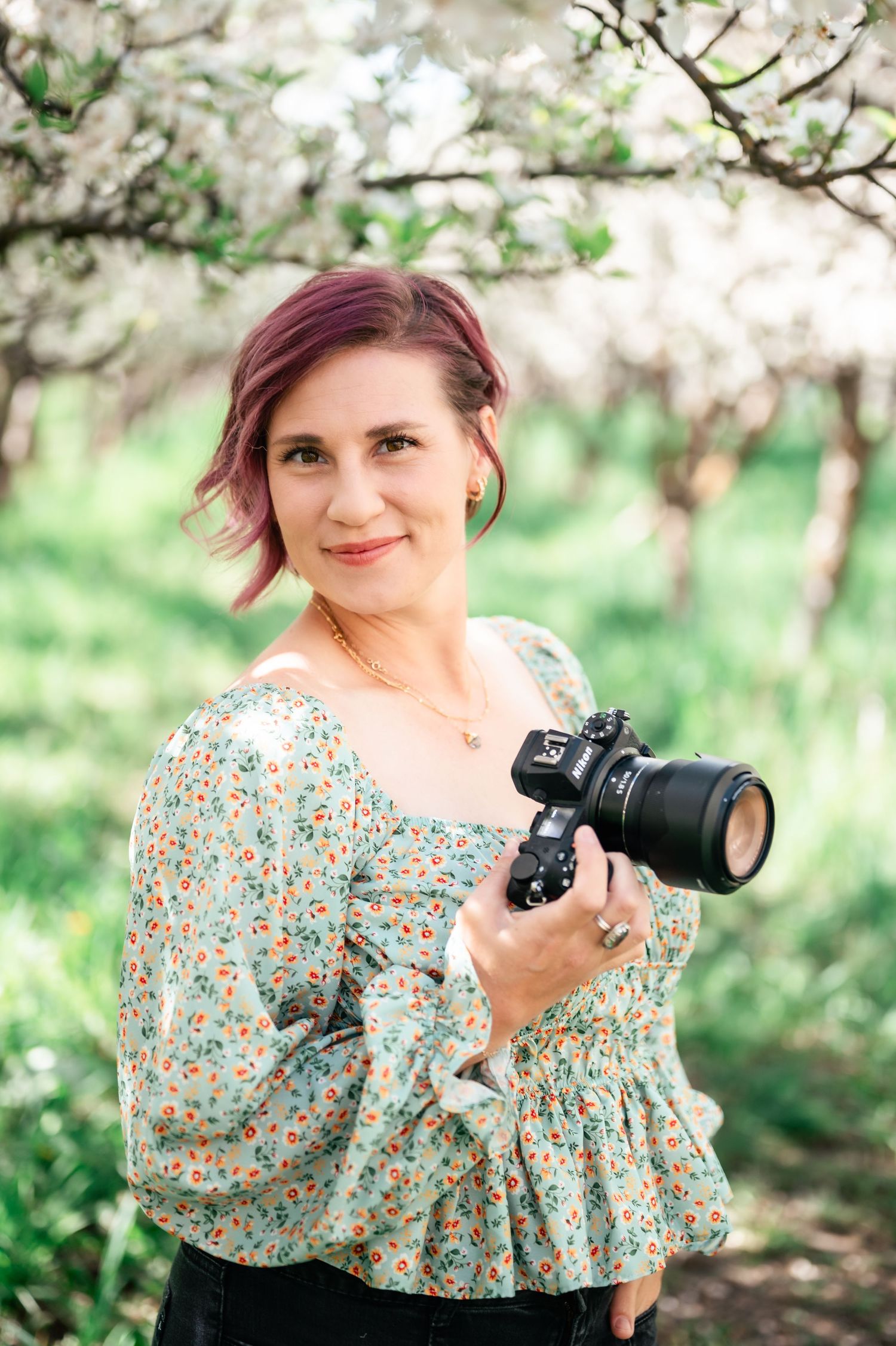 Photographer in floral patterned blouse holds professional camera while standing in blooming spring orchard.