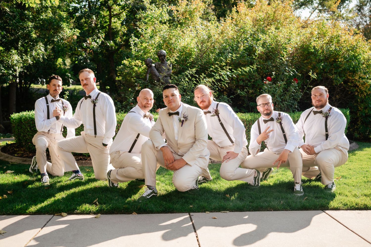 A wedding party wearing cream-colored suits poses casually on grass in a sunny garden setting.