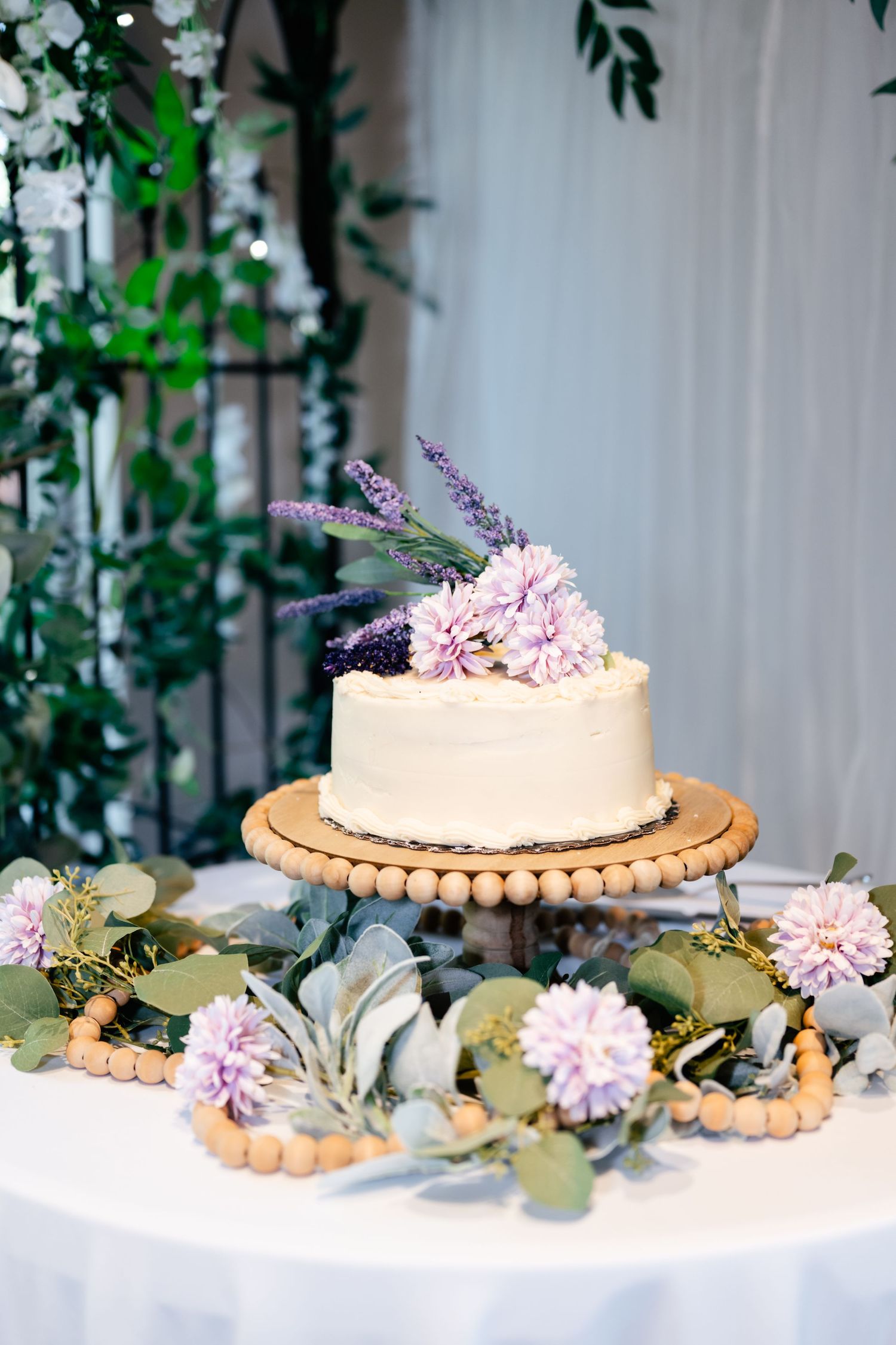 Simple white wedding cake decorated with pink carnations and lavender sprigs on a wooden stand with eucalyptus leaves.