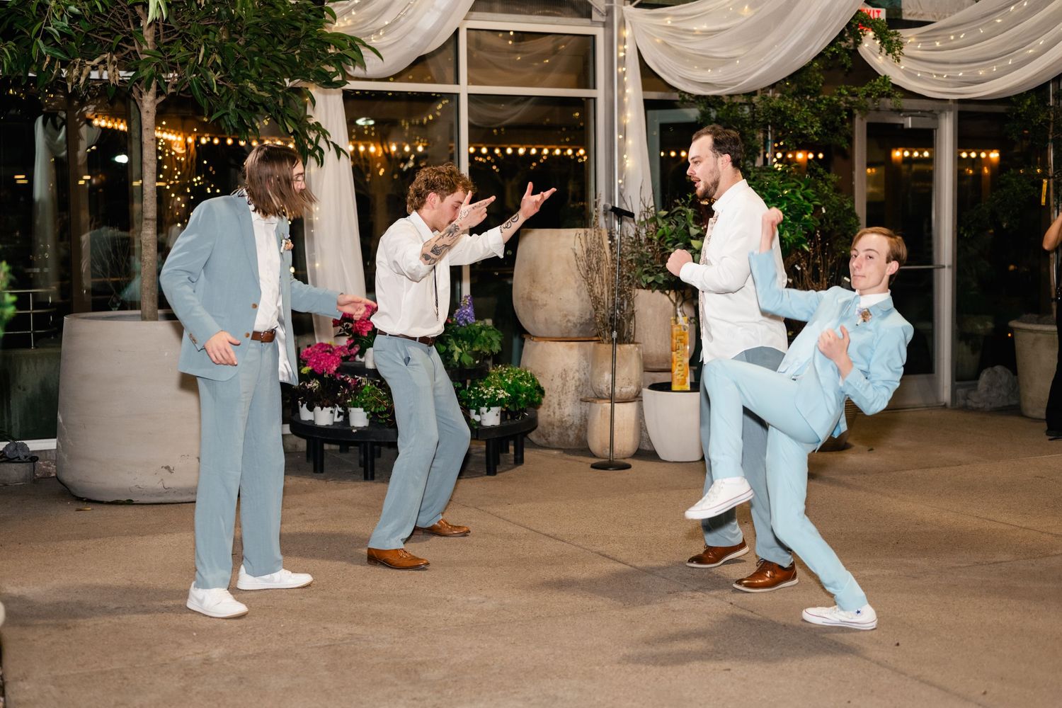 Group of wedding guests performing synchronized dance moves on an outdoor patio with string lights and drapes.