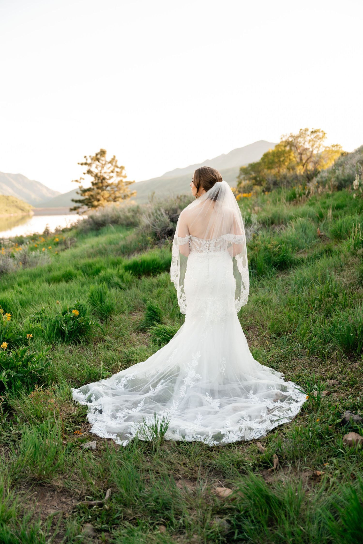 A bride in a stunning lace wedding gown with long train stands on a hillside overlooking a scenic mountain lake.