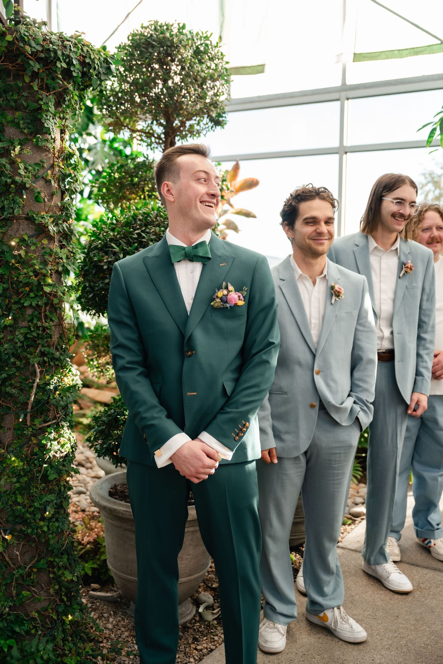 Groomsmen stand together at a greenhouse wedding venue, wearing green and gray suits with floral boutonnieres.