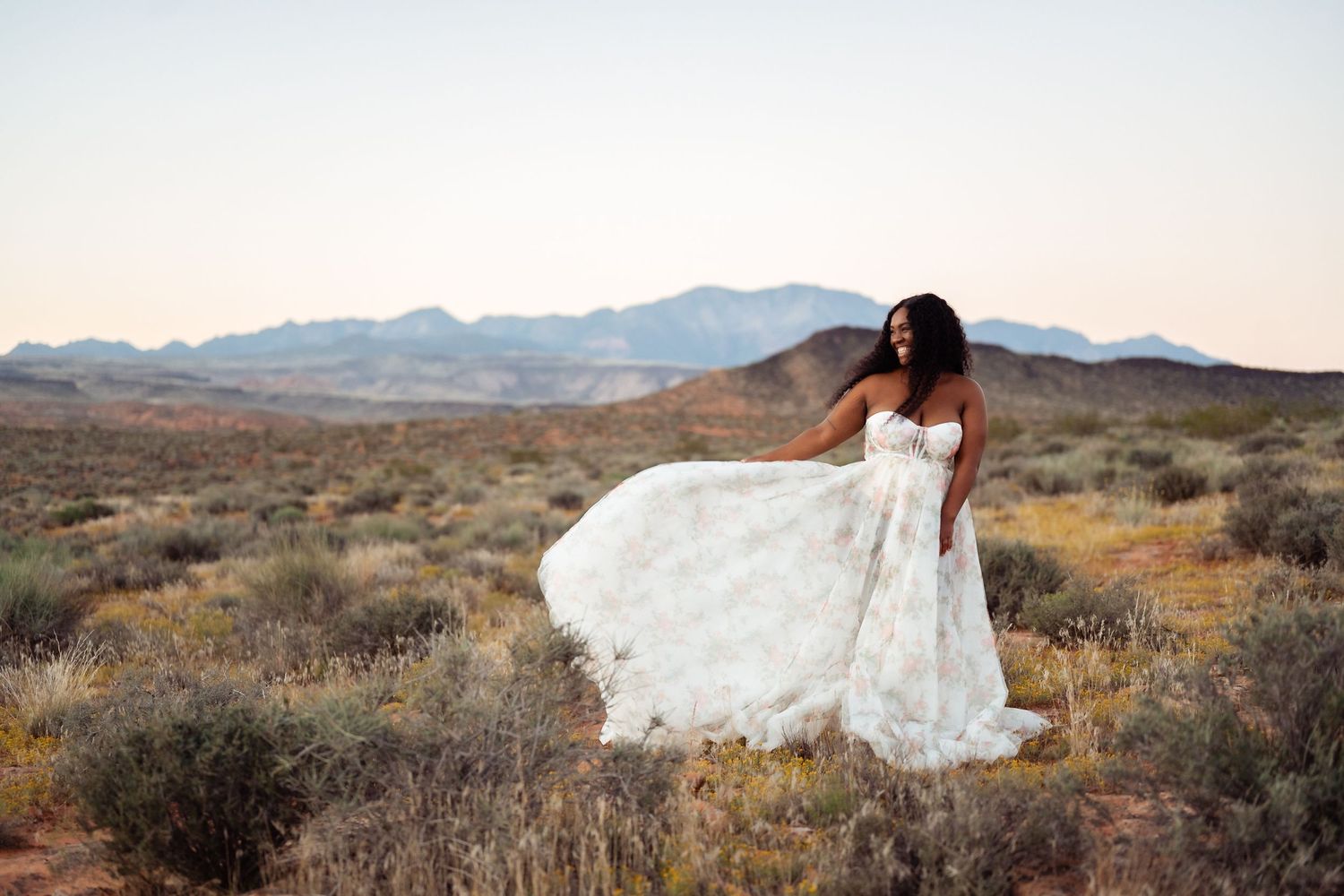 A white wedding dress flows elegantly across a desert landscape with mountains and brush in the background at sunset. Southern Utah Wedding.