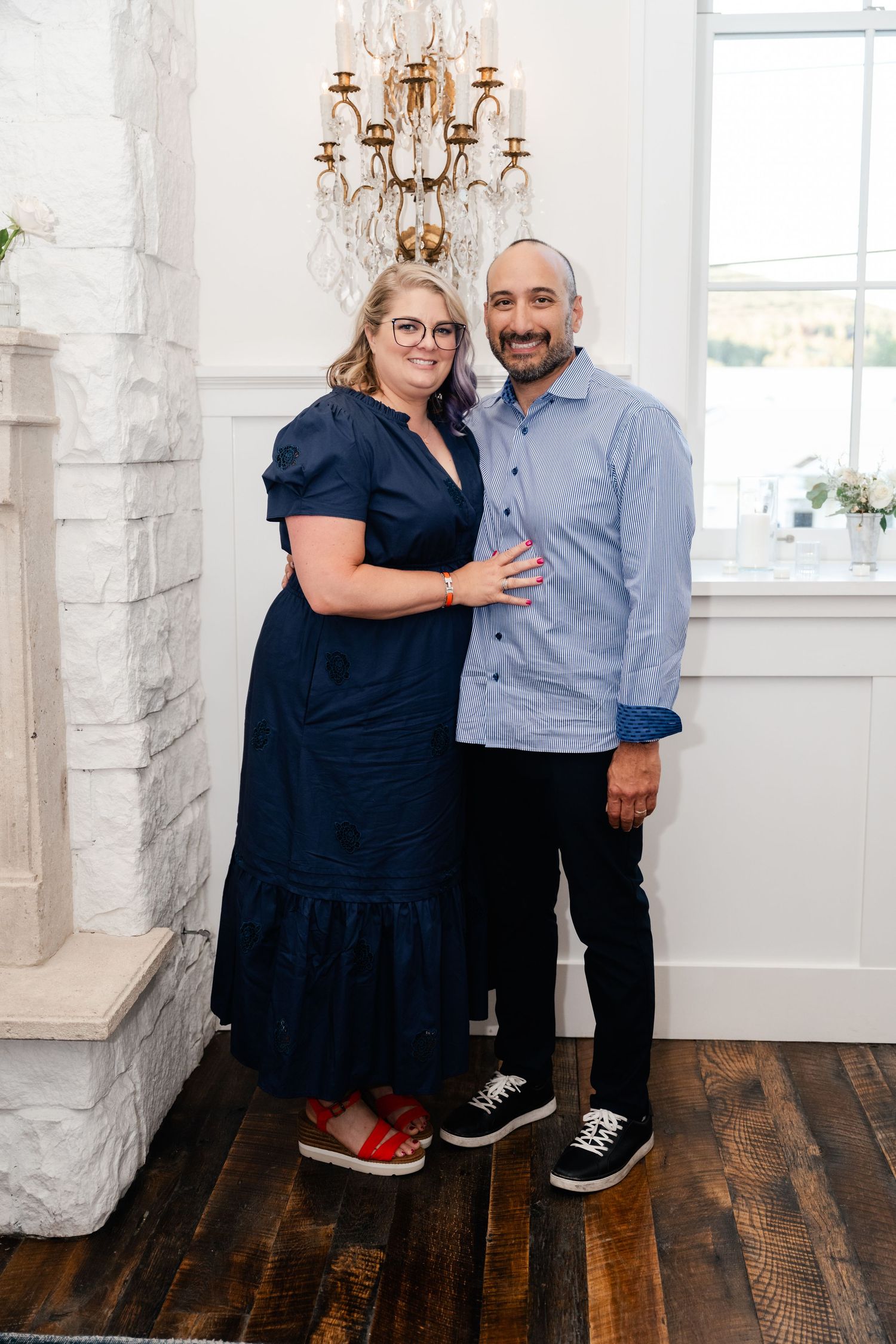 A couple poses together in a bright white room with elegant chandeliers and hardwood flooring.