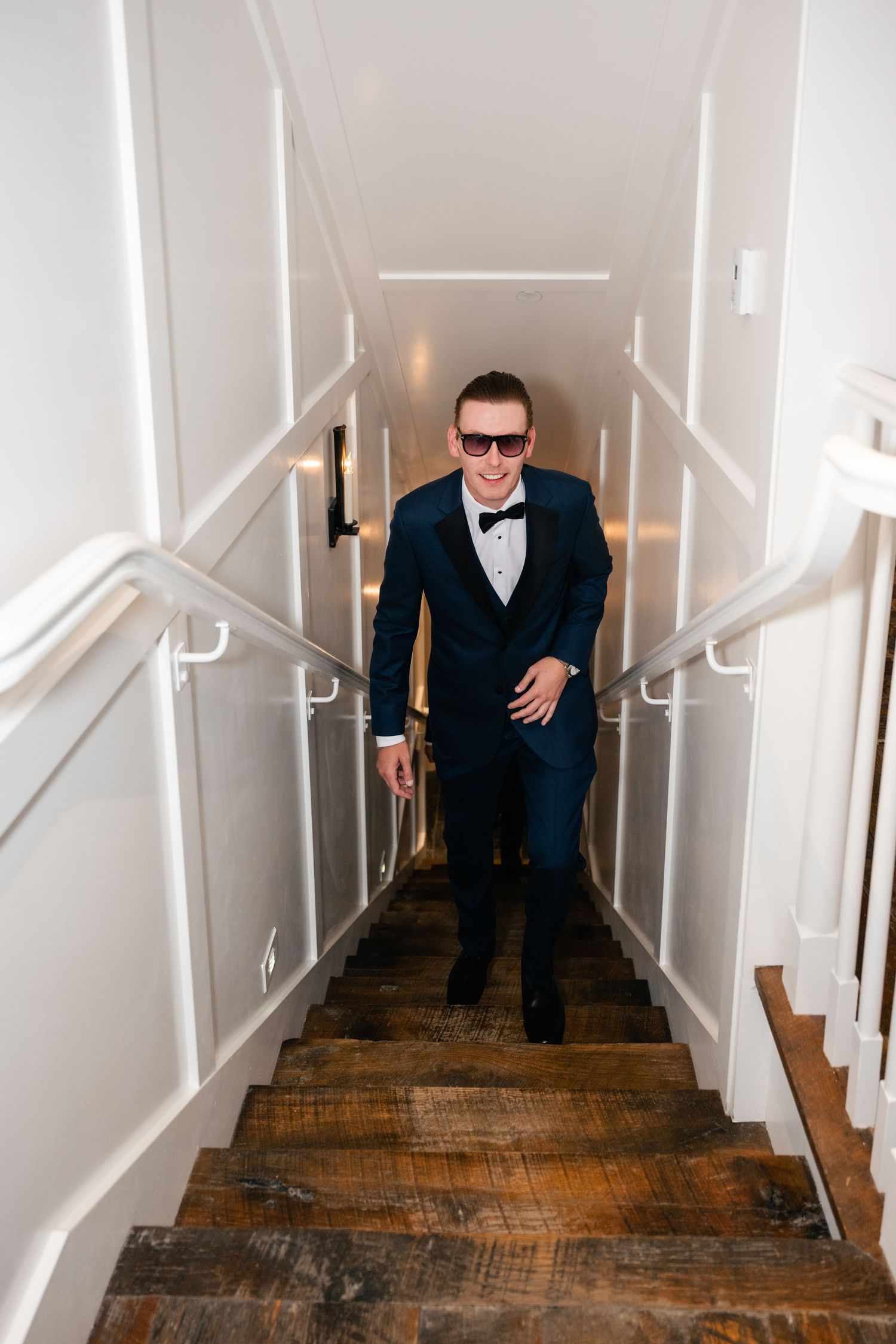 A sequence showing someone in a dark suit ascending wooden stairs with white walls and handrails.