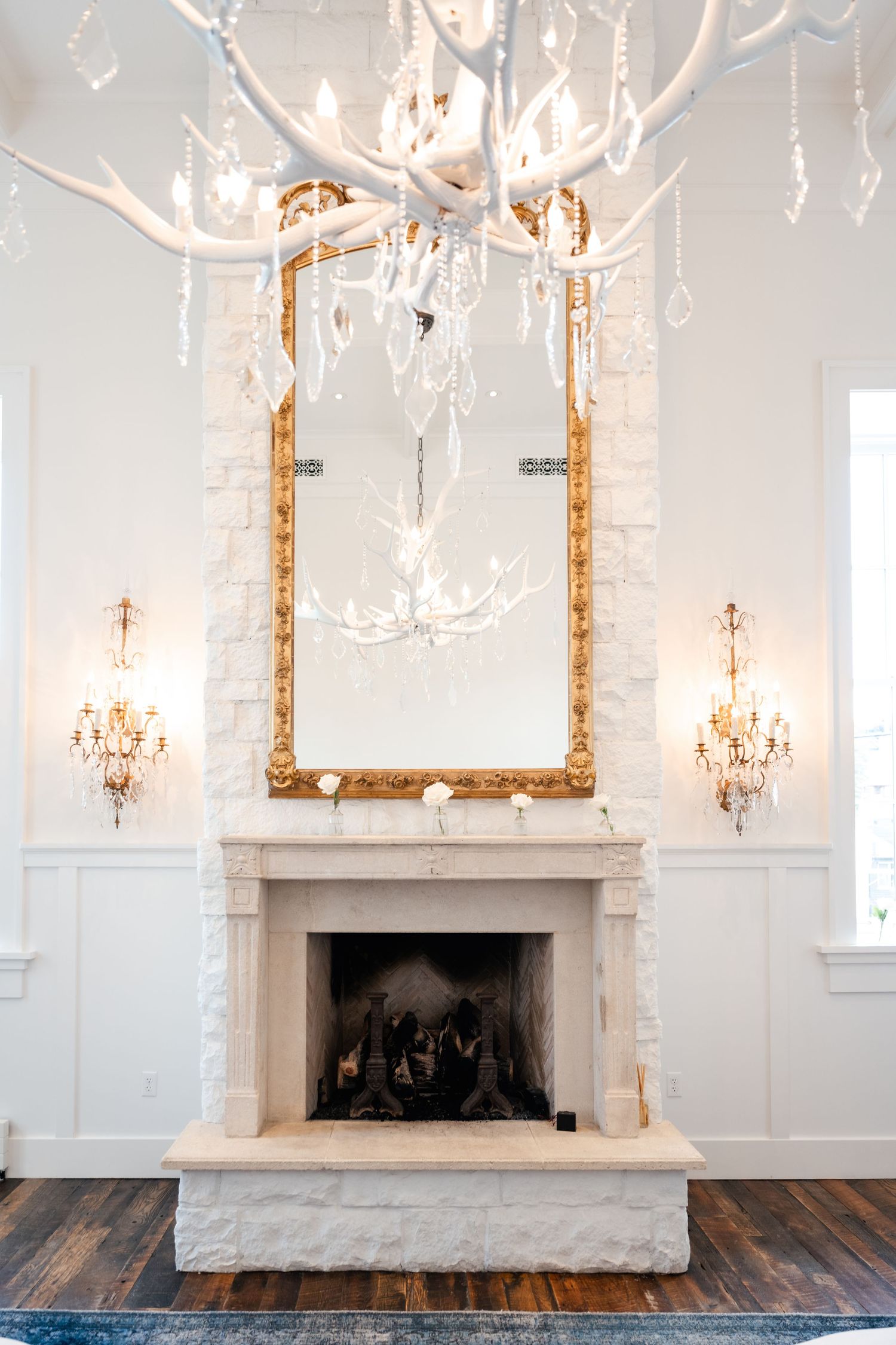 Elegant white fireplace with ornate gold mirror and antler chandelier in a luxurious room with hardwood floors.