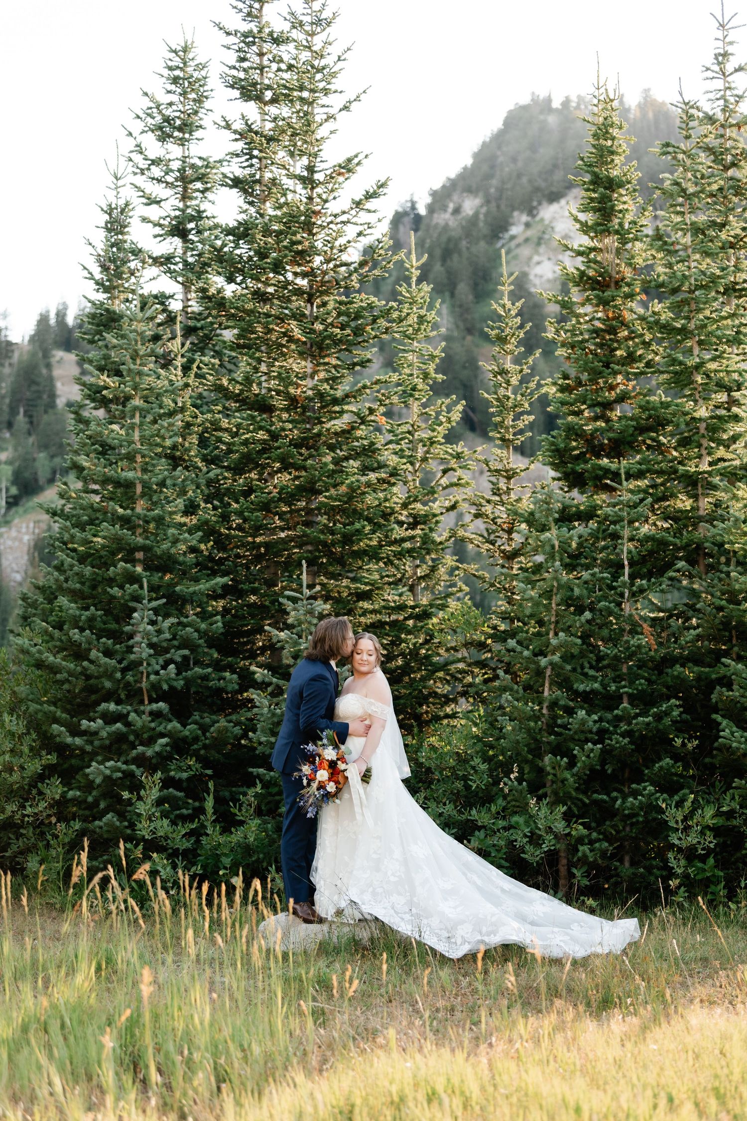 Couple shares romantic moments in a mountain meadow surrounded by evergreen trees at sunset. Utah Mountain Wedding Photographer.