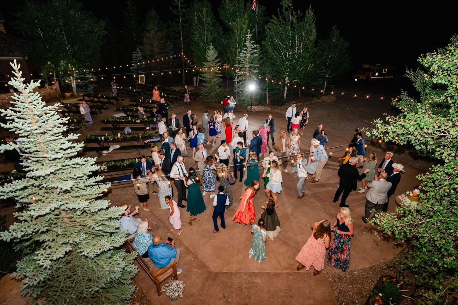 People dancing and socializing at an outdoor nighttime event with string lights and evergreen trees.