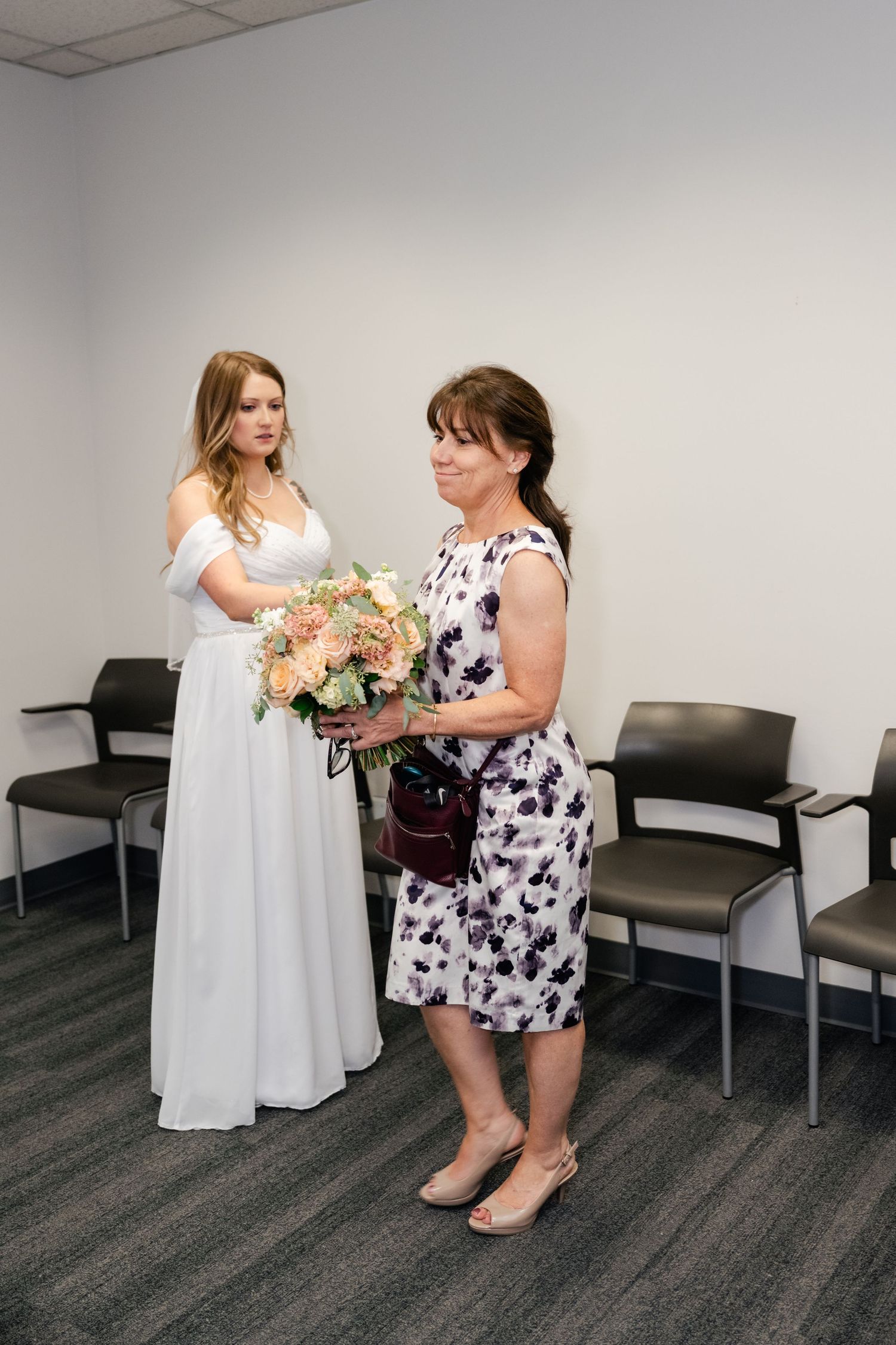 Two women embrace emotionally in a waiting room, one wearing a white dress and another in a floral print dress.
