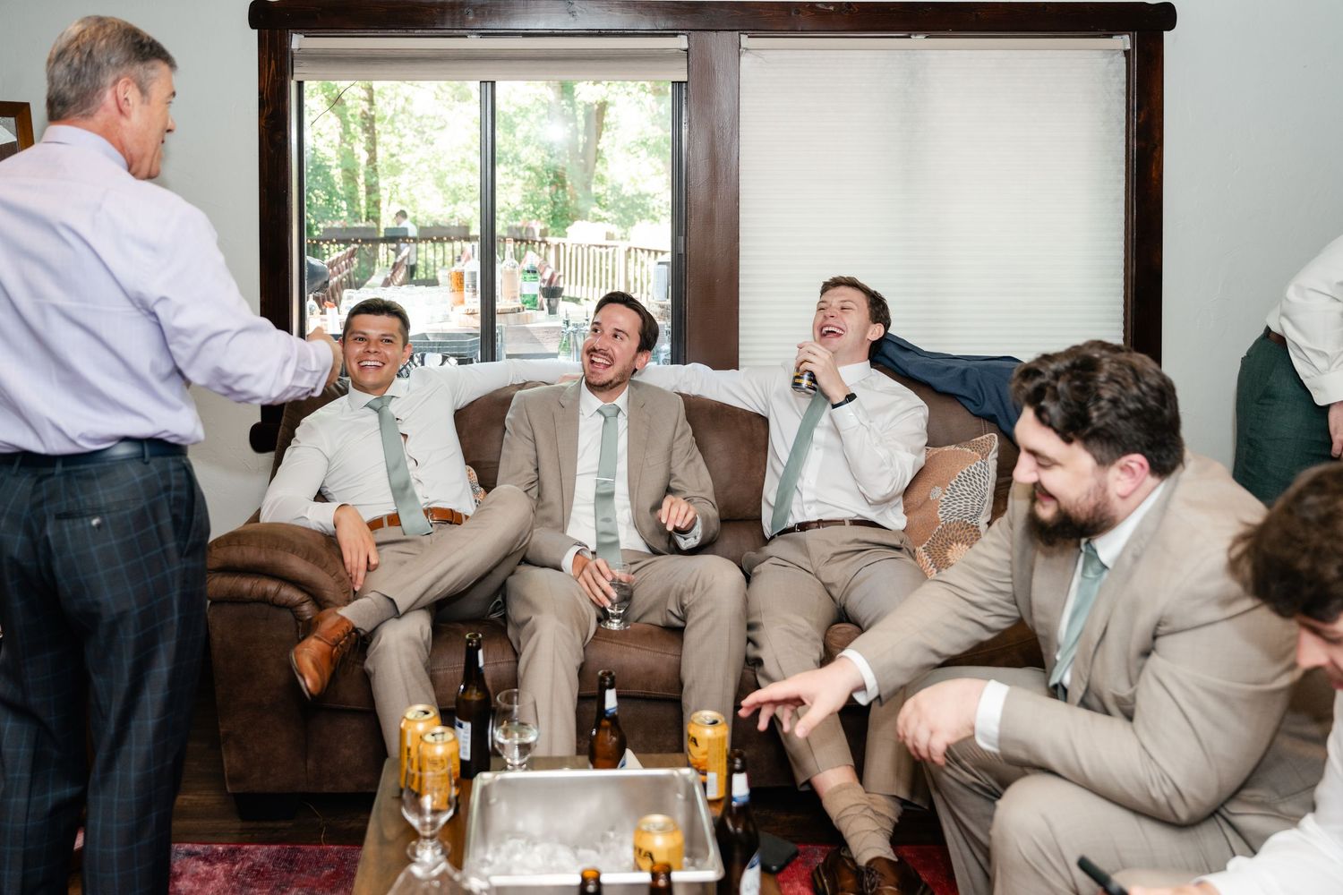 A group in light suits laughs together while relaxing on leather furniture at a social gathering. Park City Wedding Photographer.