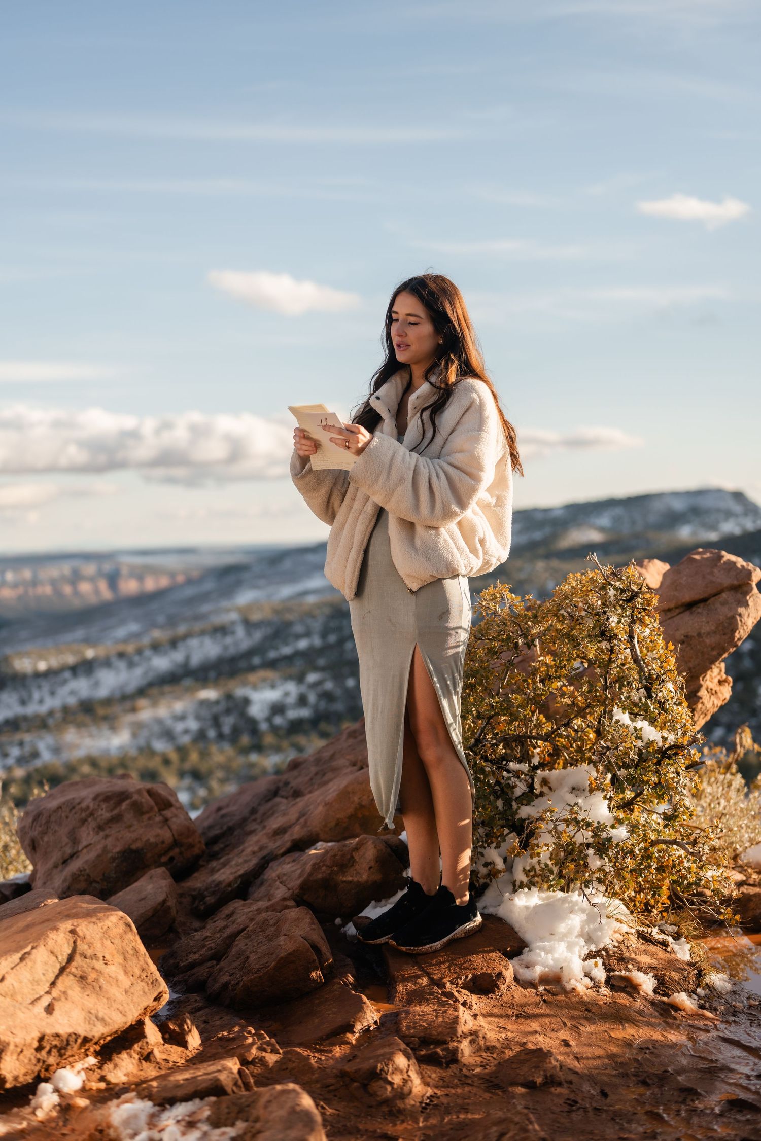 Person in cream sweater reading on red rocks with mountain views.