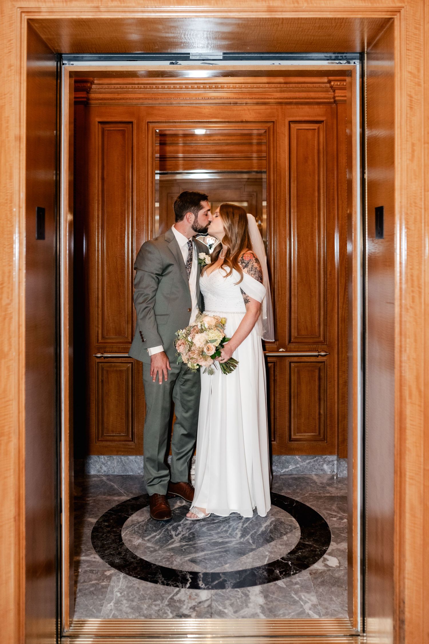 Three connected mirror panels reflect a romantic couple in formal attire kissing in an elegant wood-paneled hallway.