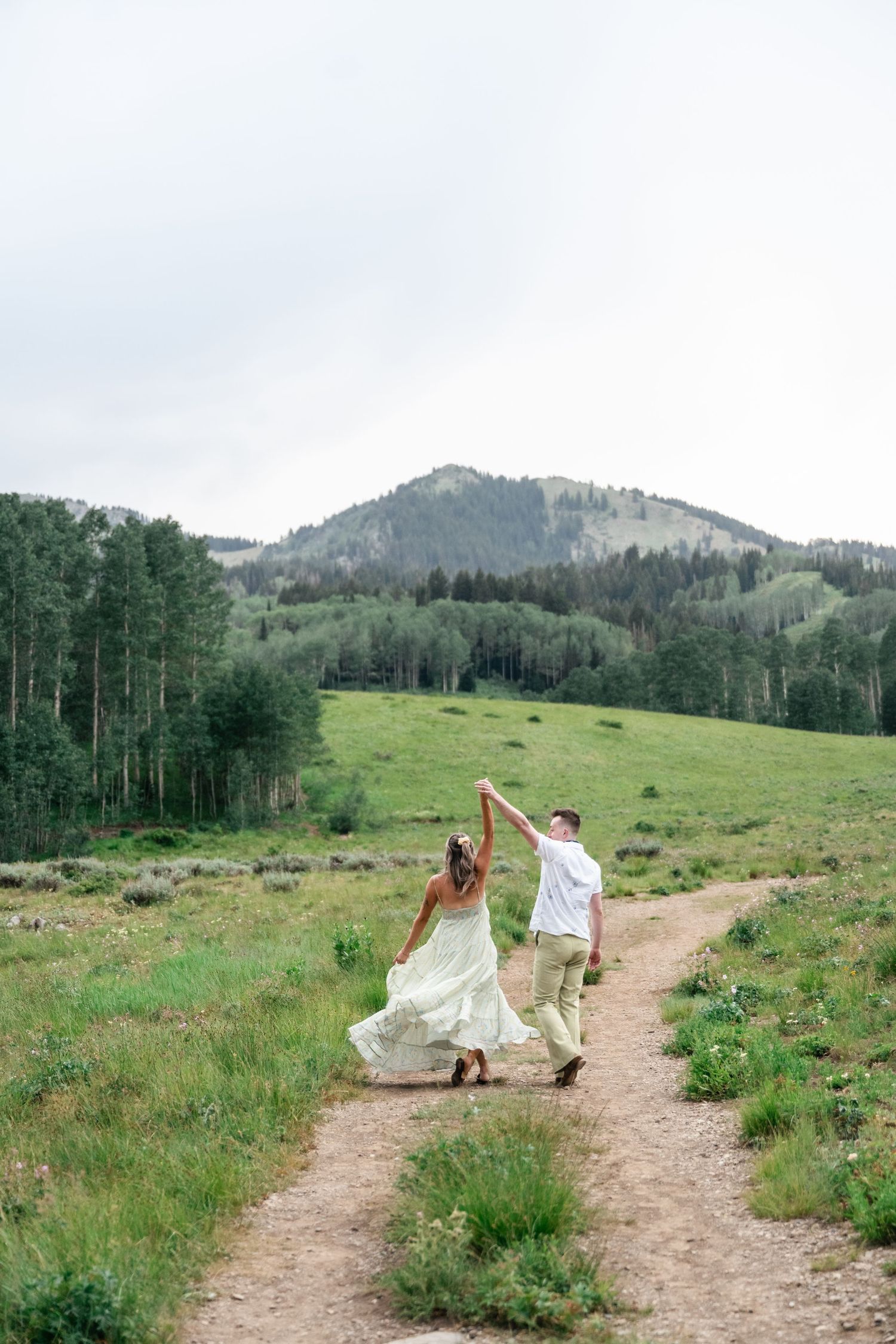 A person in flowing white fabric dances along a dirt trail through green mountain meadows with pine forests in the distance.