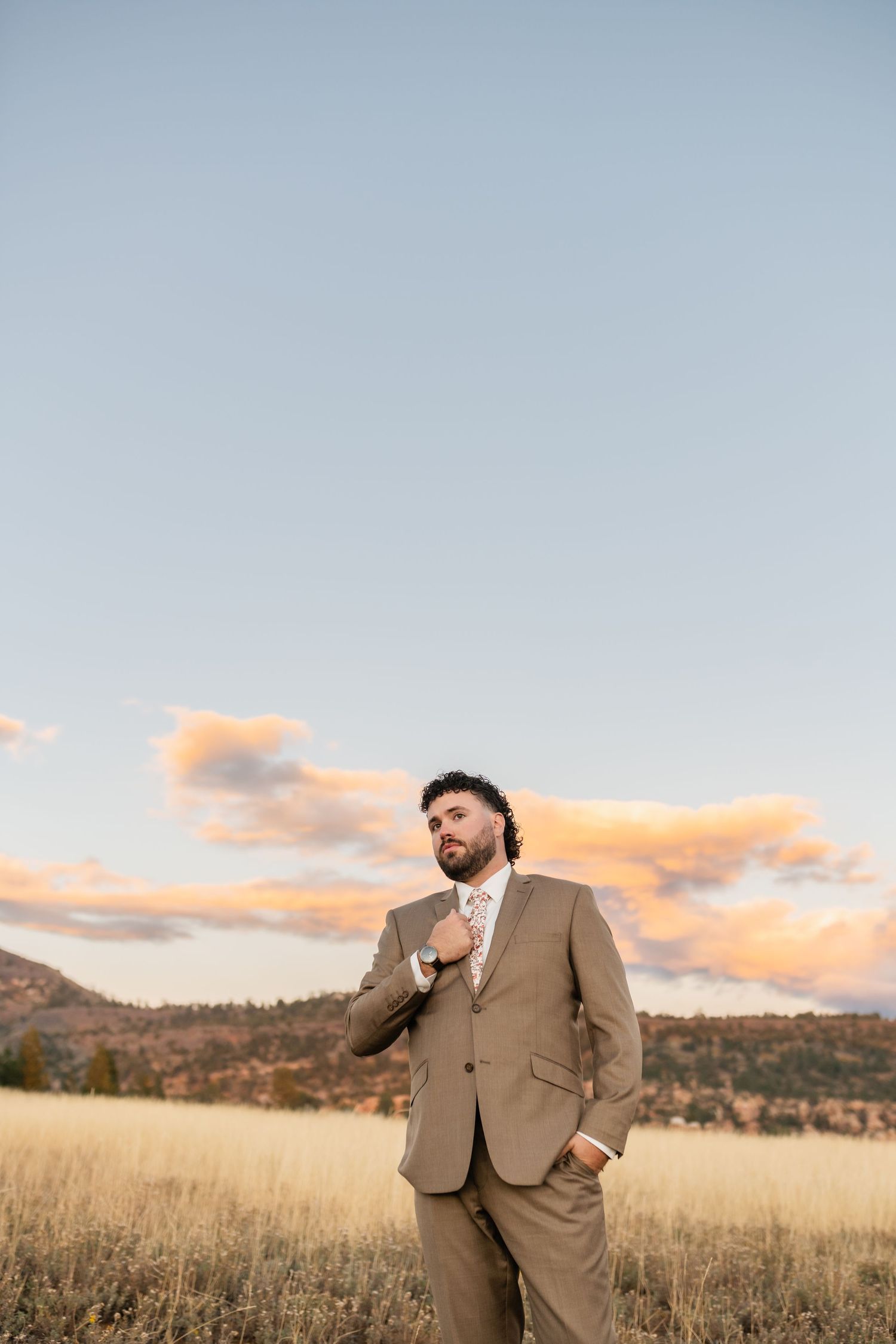 A person in formal attire poses confidently against a sunset desert backdrop.