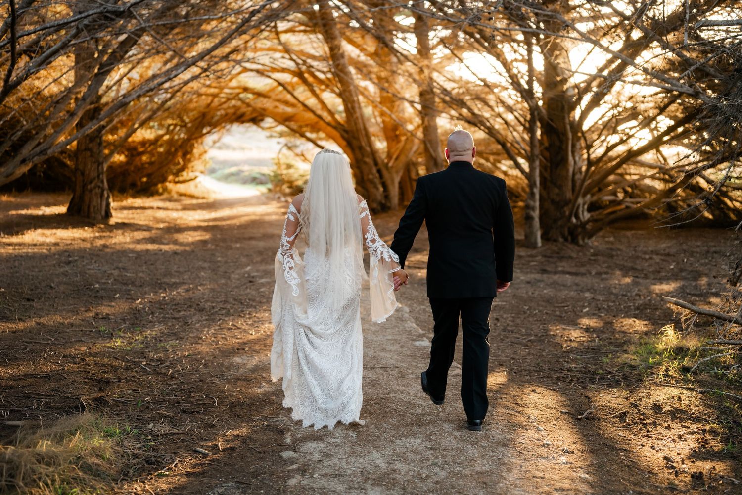 Wedding couple walking hand in hand through tree-lined path at sunset.