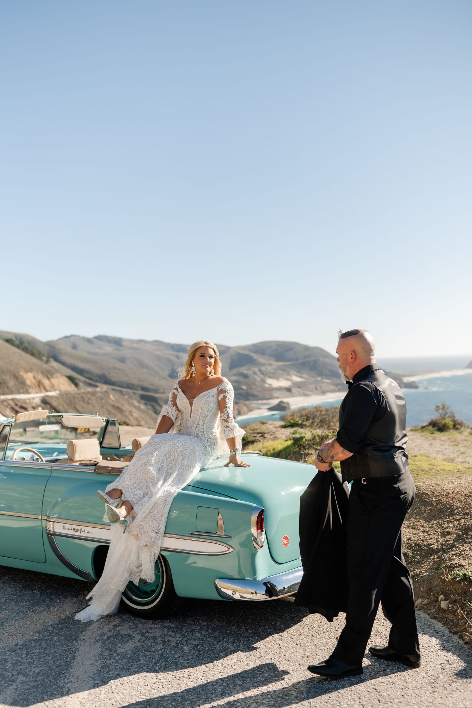 A couple poses near a turquoise vintage car with scenic mountain and water views in the background.