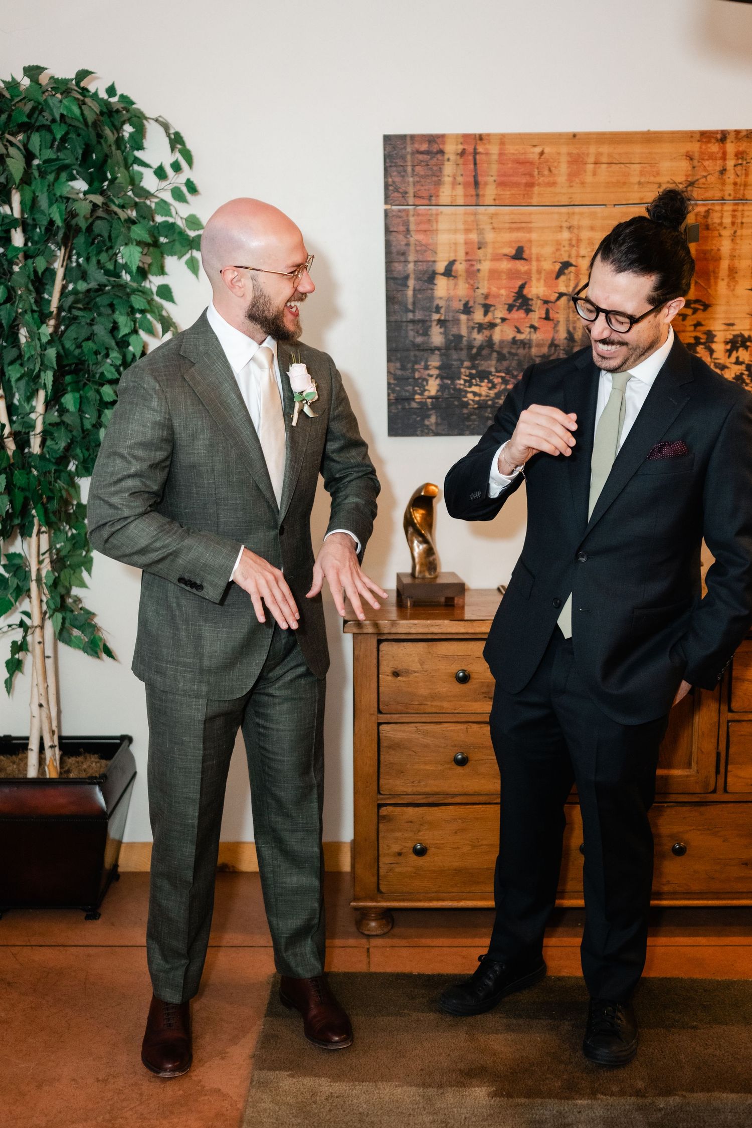 Two wedding guests in suits share a lighthearted moment together in front of a decorative wall and plant.