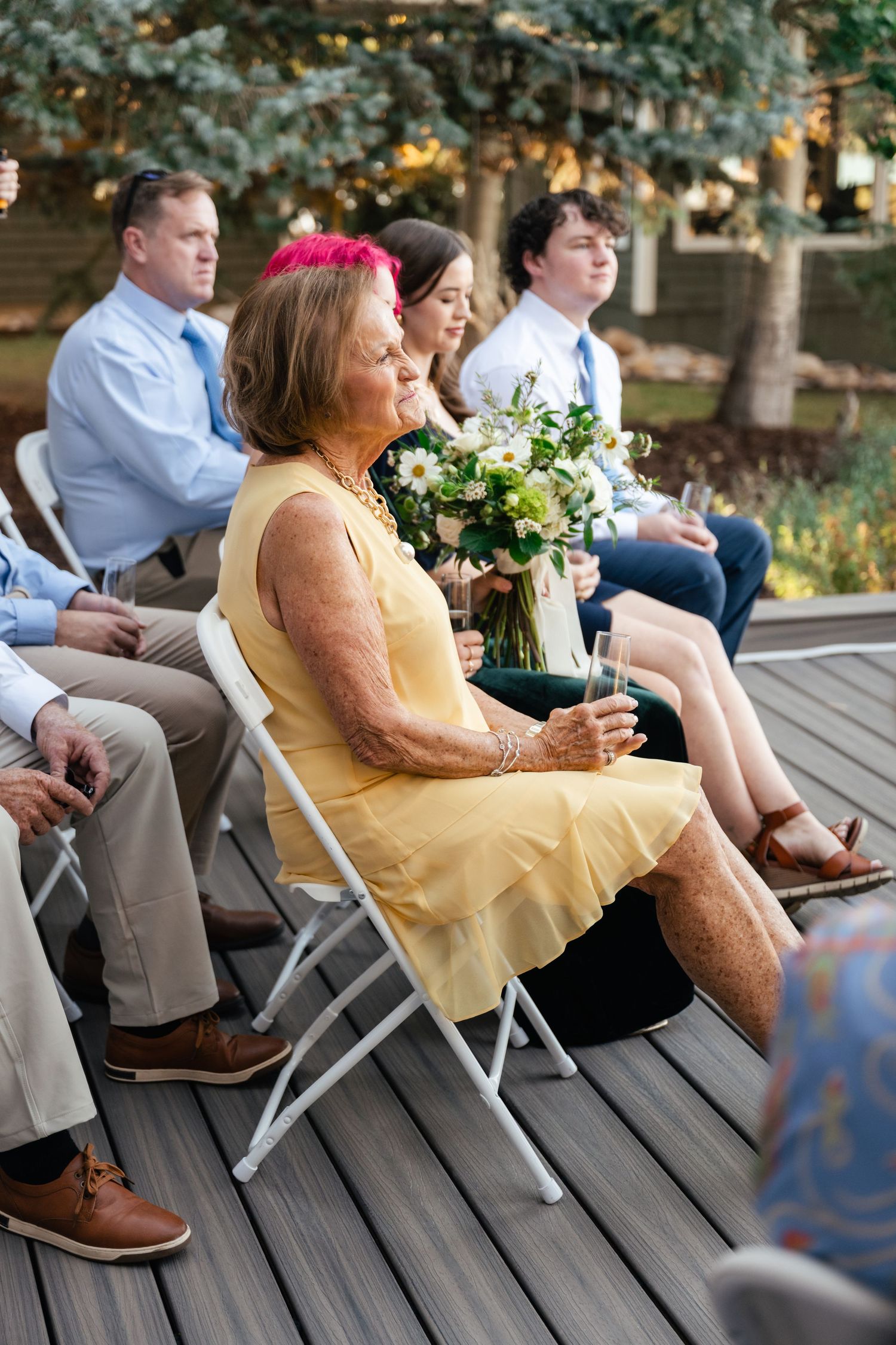 Wedding guests seated in folding chairs on a wooden deck during an outdoor ceremony.