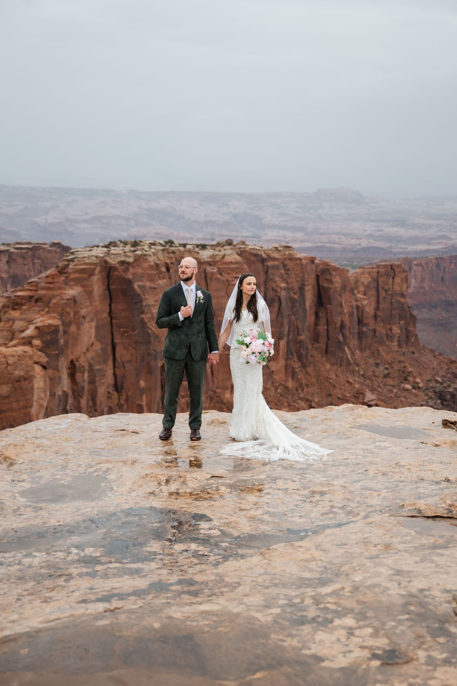 A couple celebrates their wedding day on the edge of a dramatic red rock canyon with vast desert views in the background.