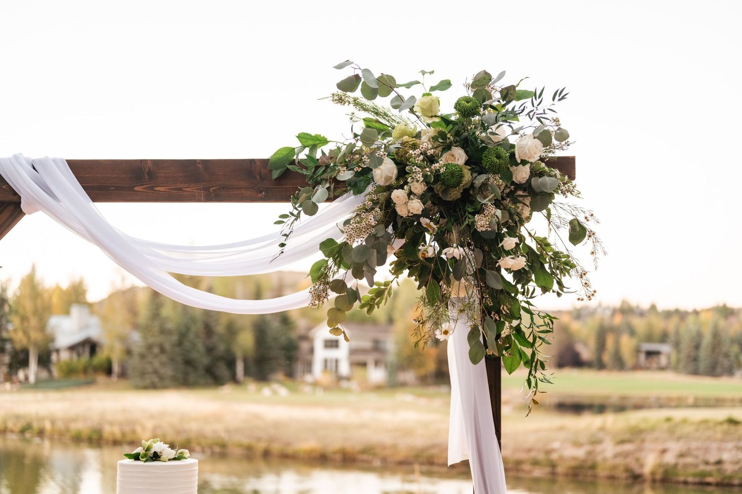 An outdoor wedding arch decorated with white fabric and floral arrangements beside a lake.