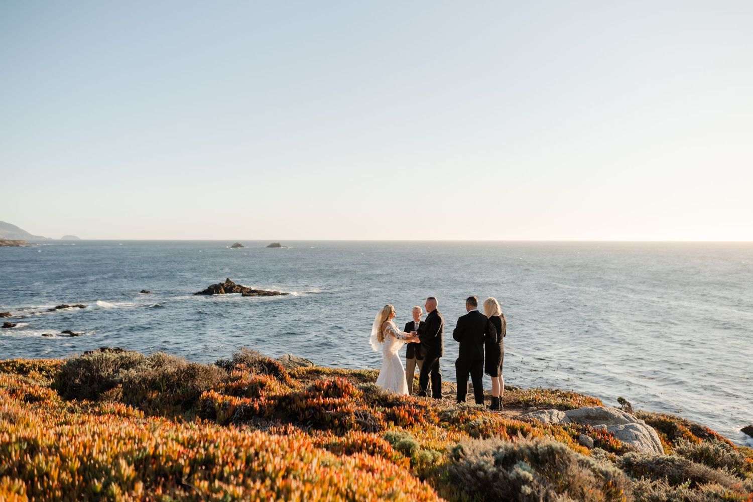 Panoramic view of wedding ceremony guests on California coastal cliffs at sunset.