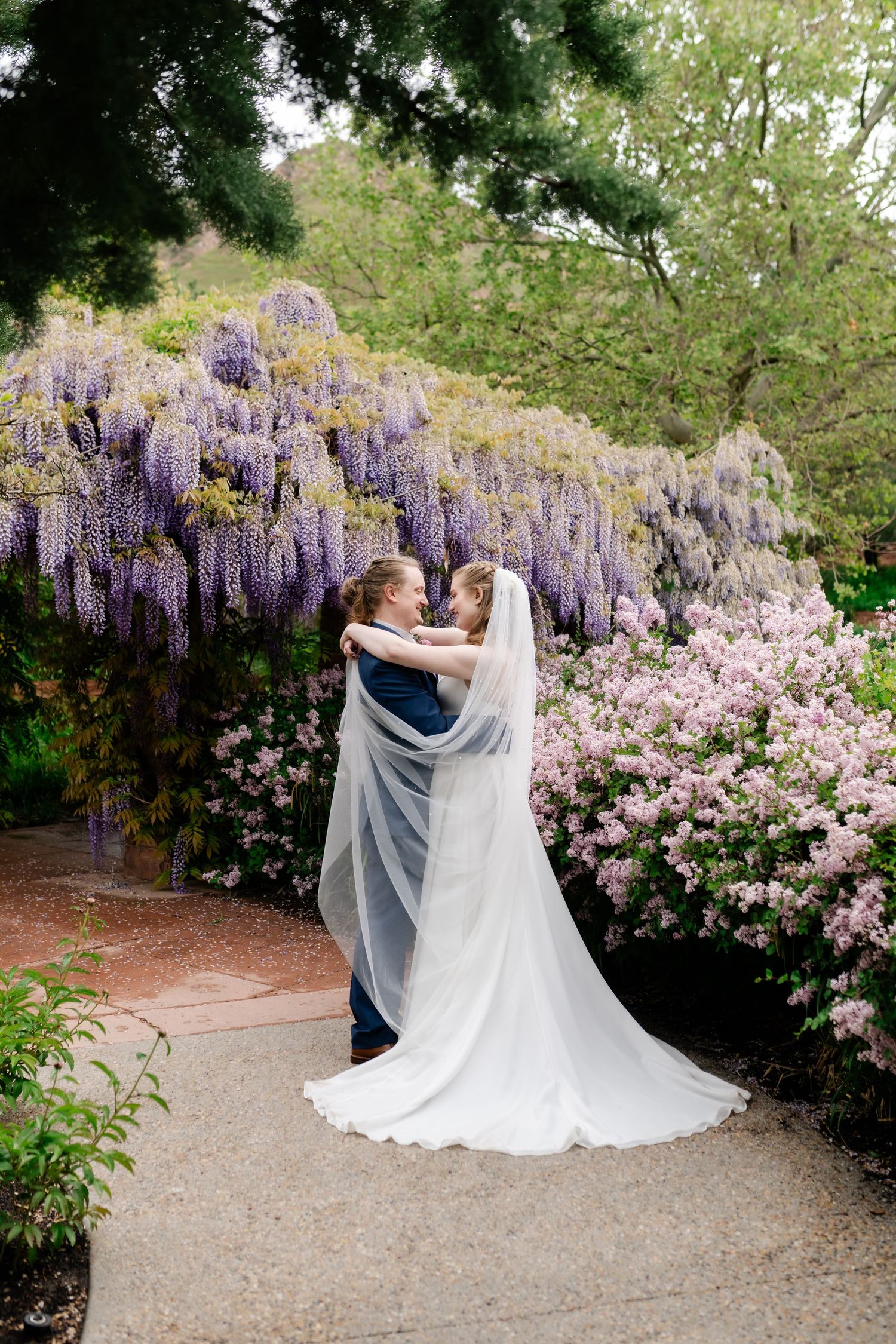 A romantic wedding moment captured under cascading purple wisteria and pink blossoms in a garden setting.