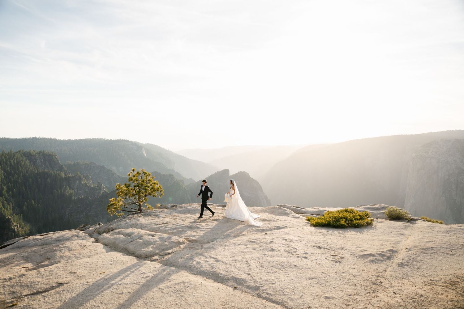 Bride and groom walk on granite cliff at Yosemite with sweeping valley views and Half Dome in the background.