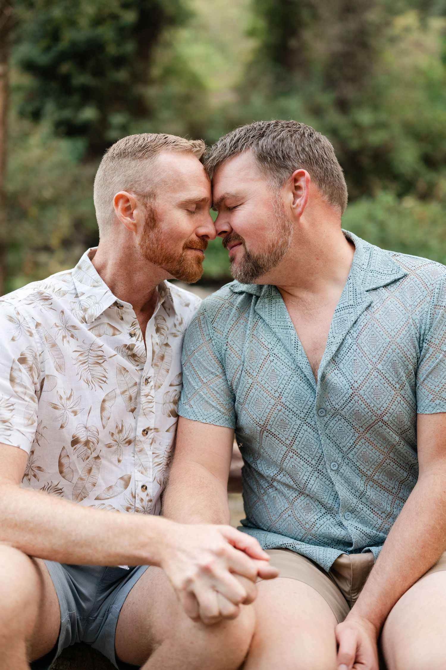 Two men holding hands and touching foreheads in an intimate romantic moment outdoors surrounded by lush green trees. Gay engagement photos.