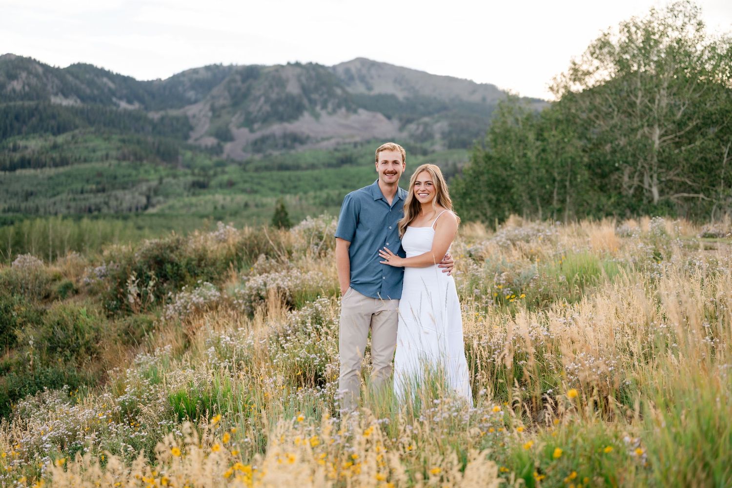 Couple smiling together in a mountain meadow with wildflowers, trees, and scenic hills during their Park City engagements.