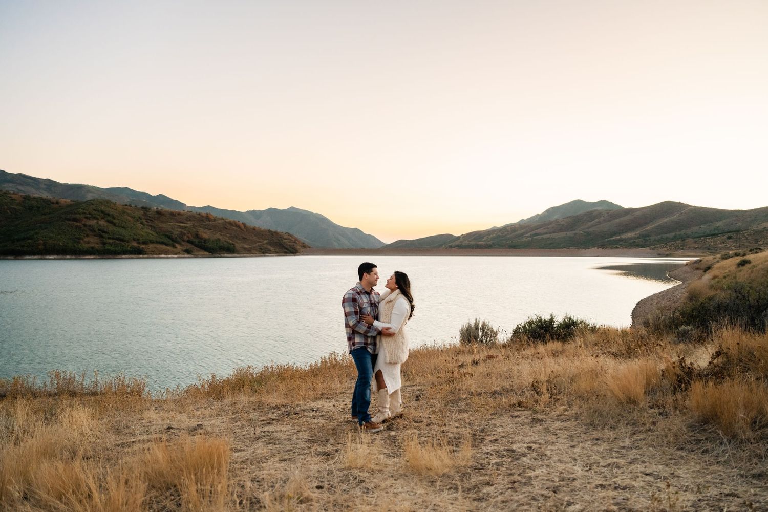 A couple embraces on a hilltop overlooking a serene mountain lake at sunset in a scenic wilderness landscape.