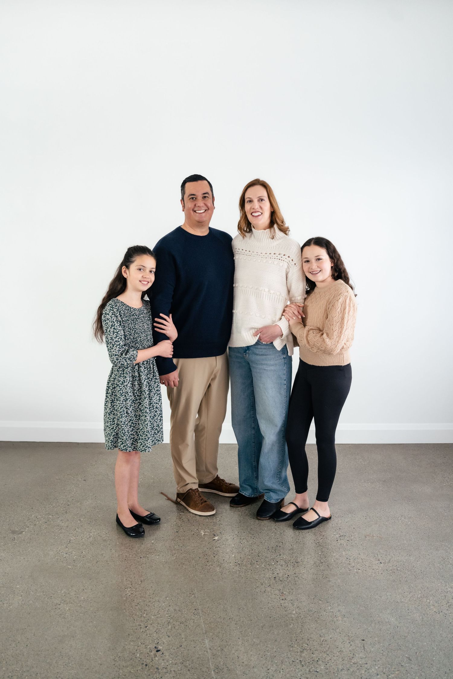 A family poses together in casual clothing against a white backdrop in a minimalist studio setting.