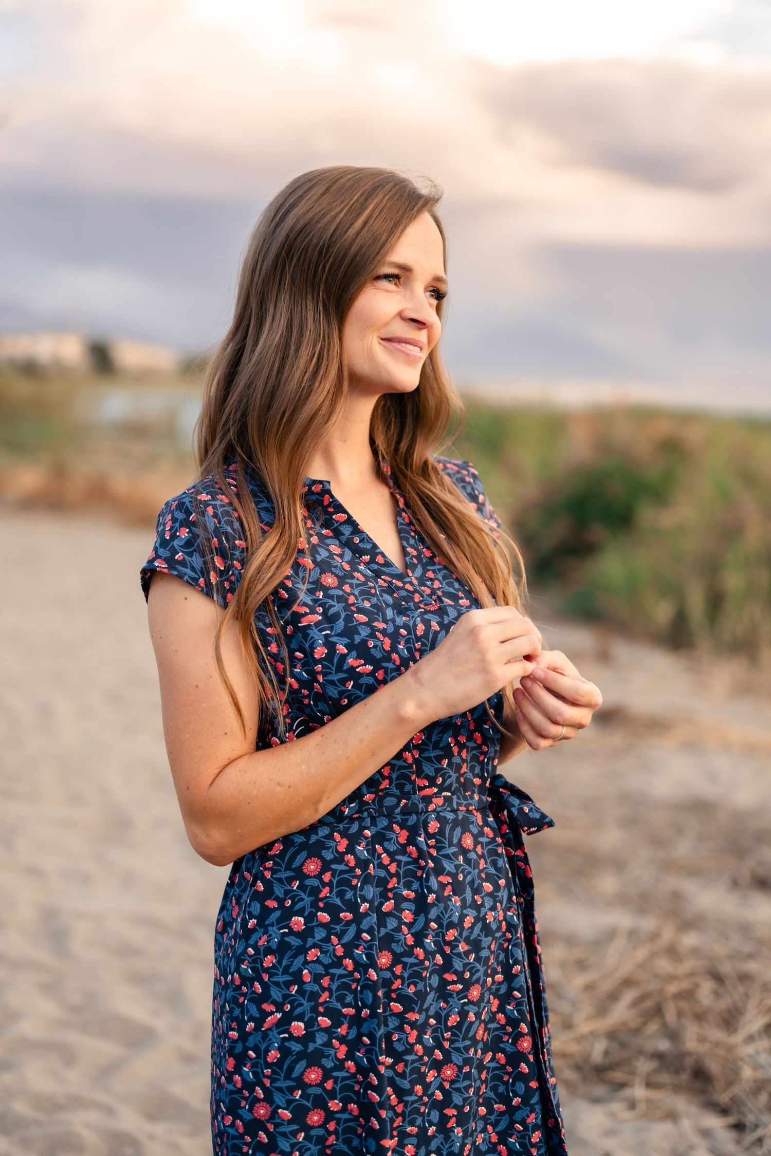 A person in a navy floral dress stands on a sandy beach during golden hour.
