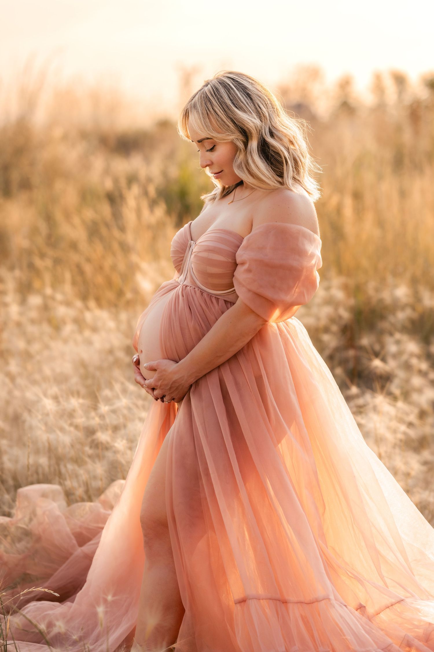 A person in a flowing peach-colored tulle gown poses gracefully in a sun-lit field during golden hour.