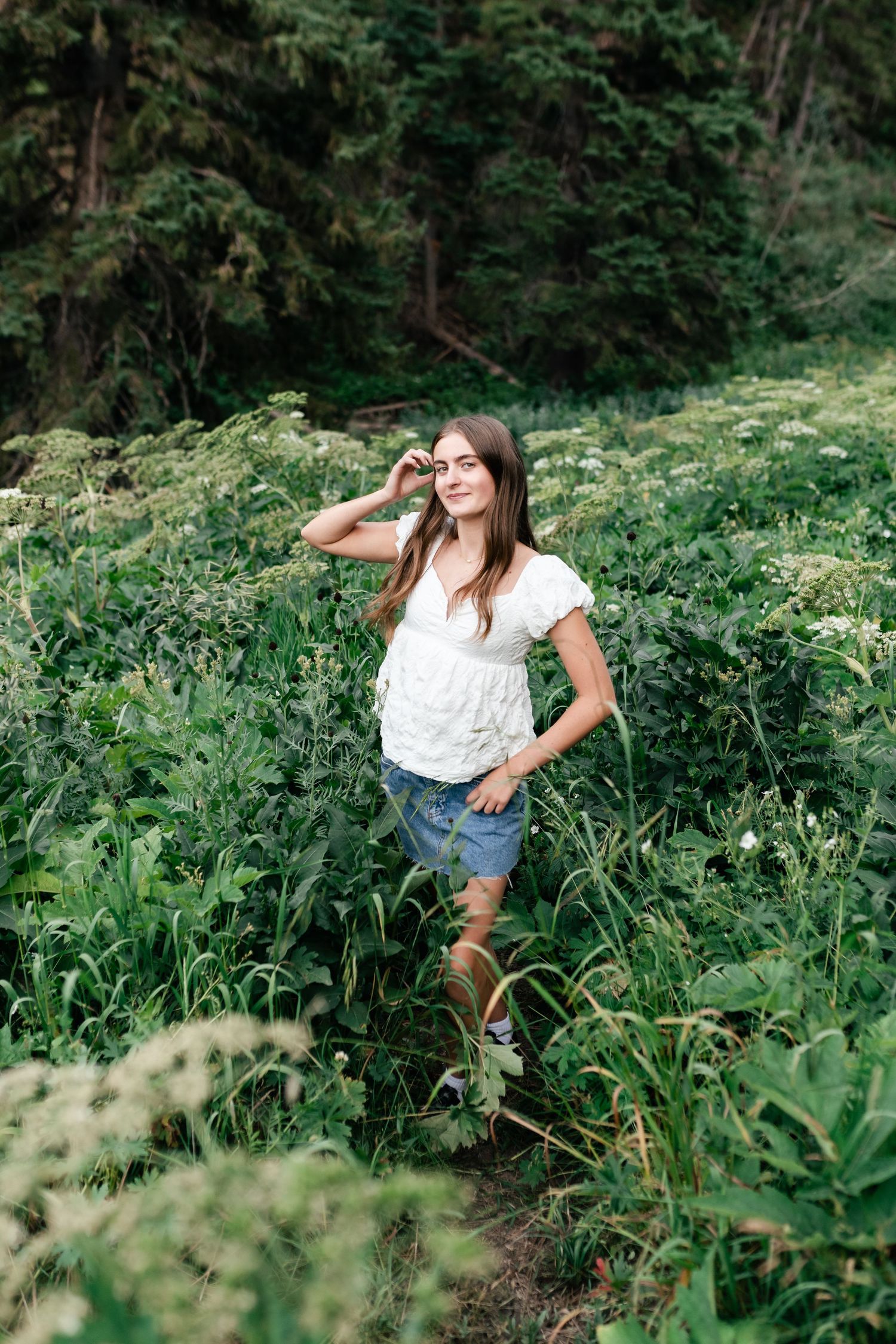 A person in a white top and denim shorts poses playfully among lush green foliage in a forest setting.
