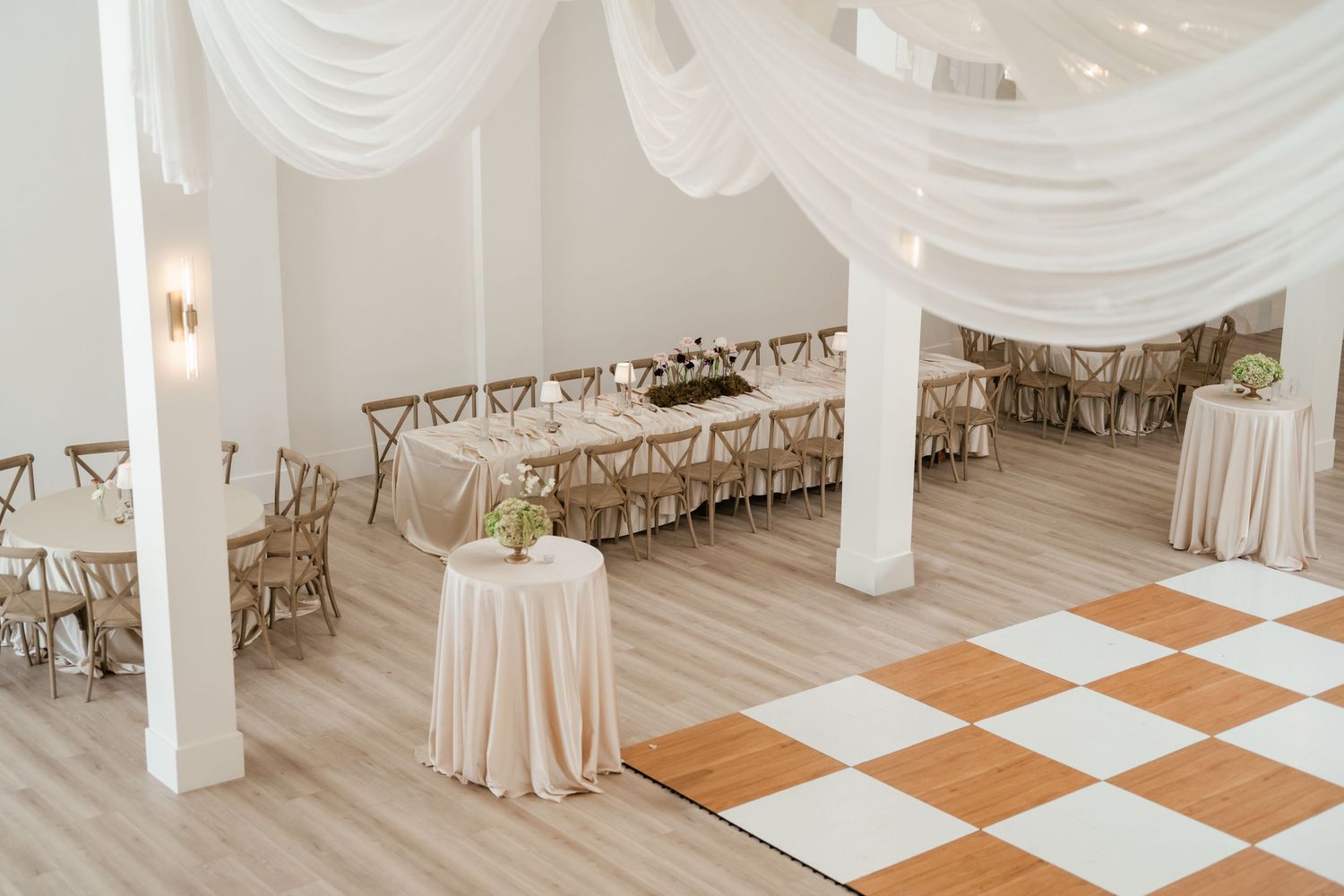 Elegant wedding reception venue with checkerboard dance floor, white draped fabric, and round tables with neutral linens. Moments on Main.