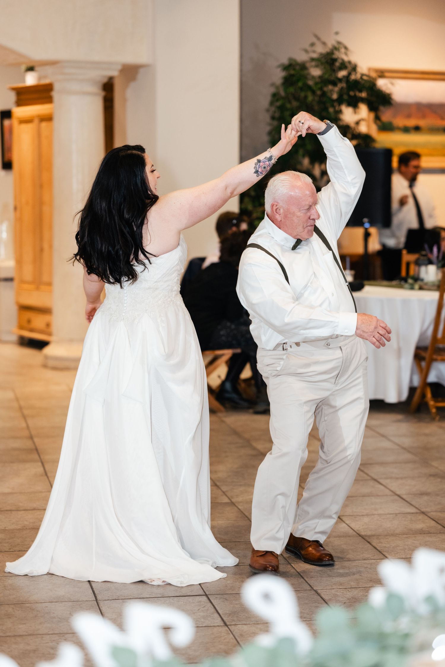 A woman in a white wedding dress twirls on the dance floor with an older gentleman in a white dress shirt.