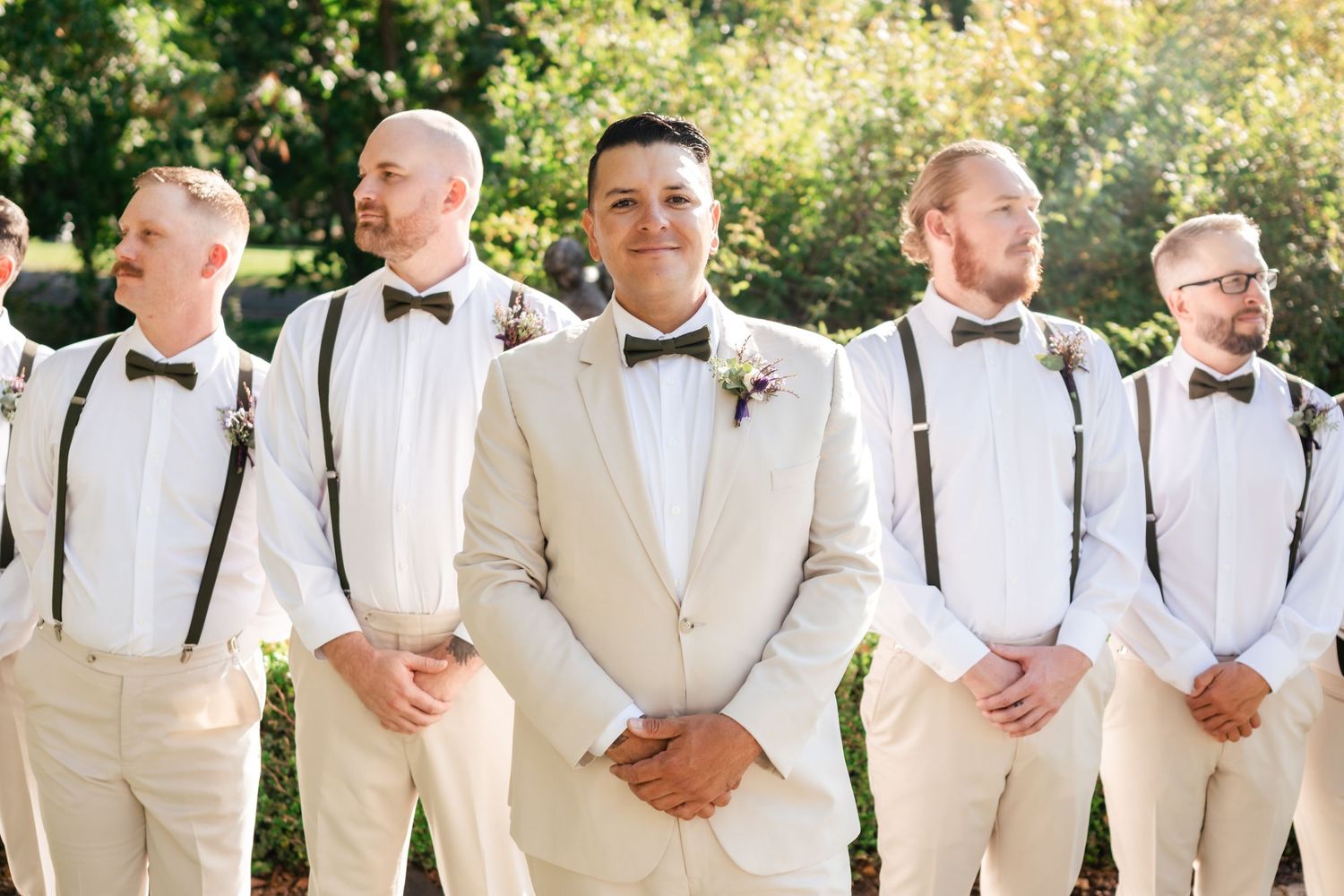 A wedding party with groomsmen wearing suspenders and bowties standing together outdoors on a sunny day.