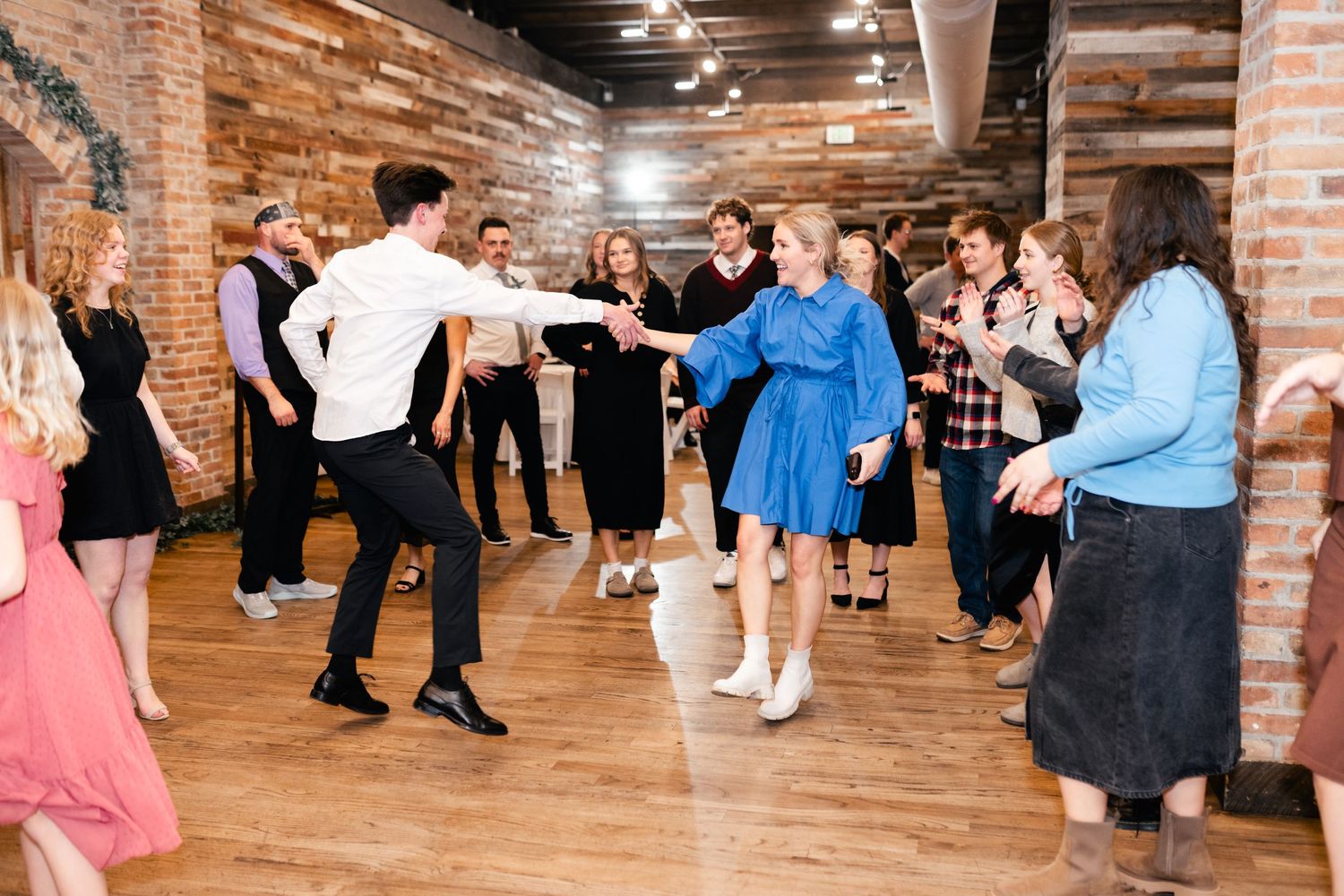 People dance in a rustic venue with exposed brick walls and hardwood flooring during a celebration.
