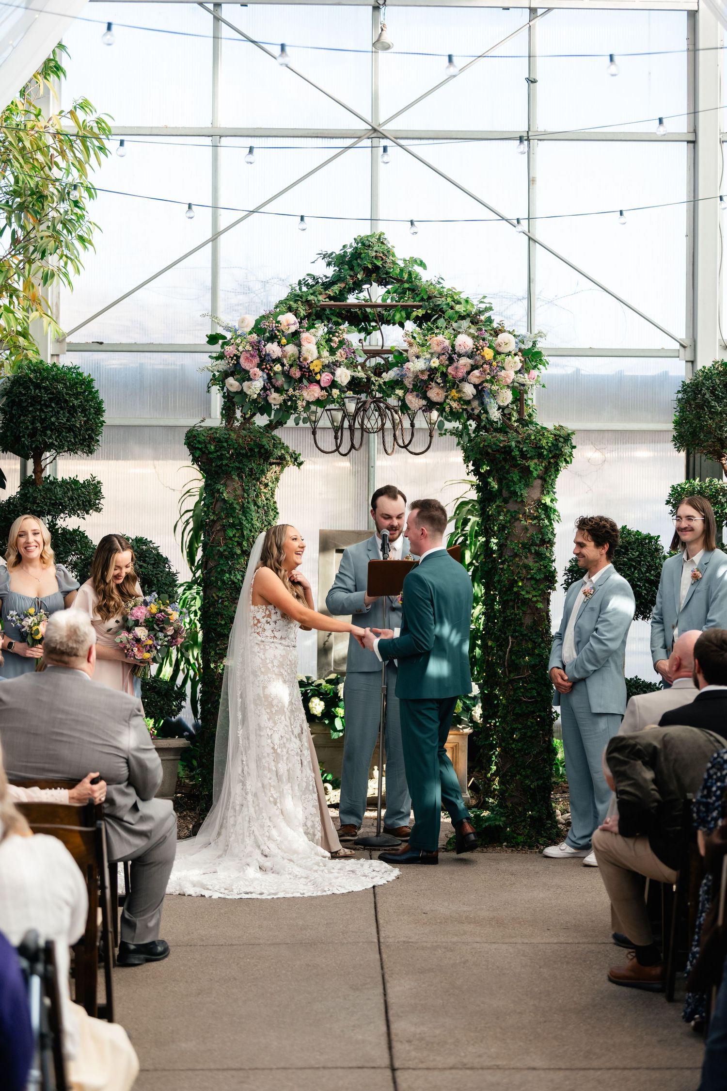 Wedding ceremony under a floral arch with string lights in greenhouse venue.