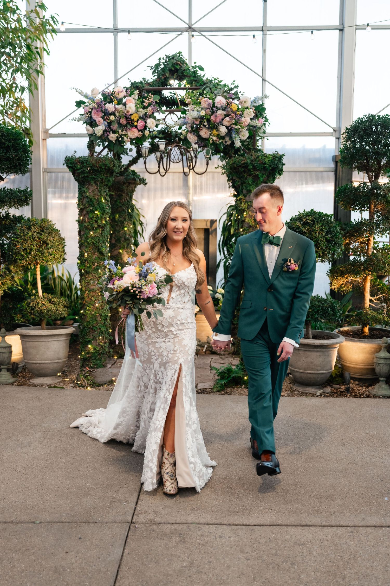 Couple walks together in greenhouse venue decorated with floral arrangements and potted plants.