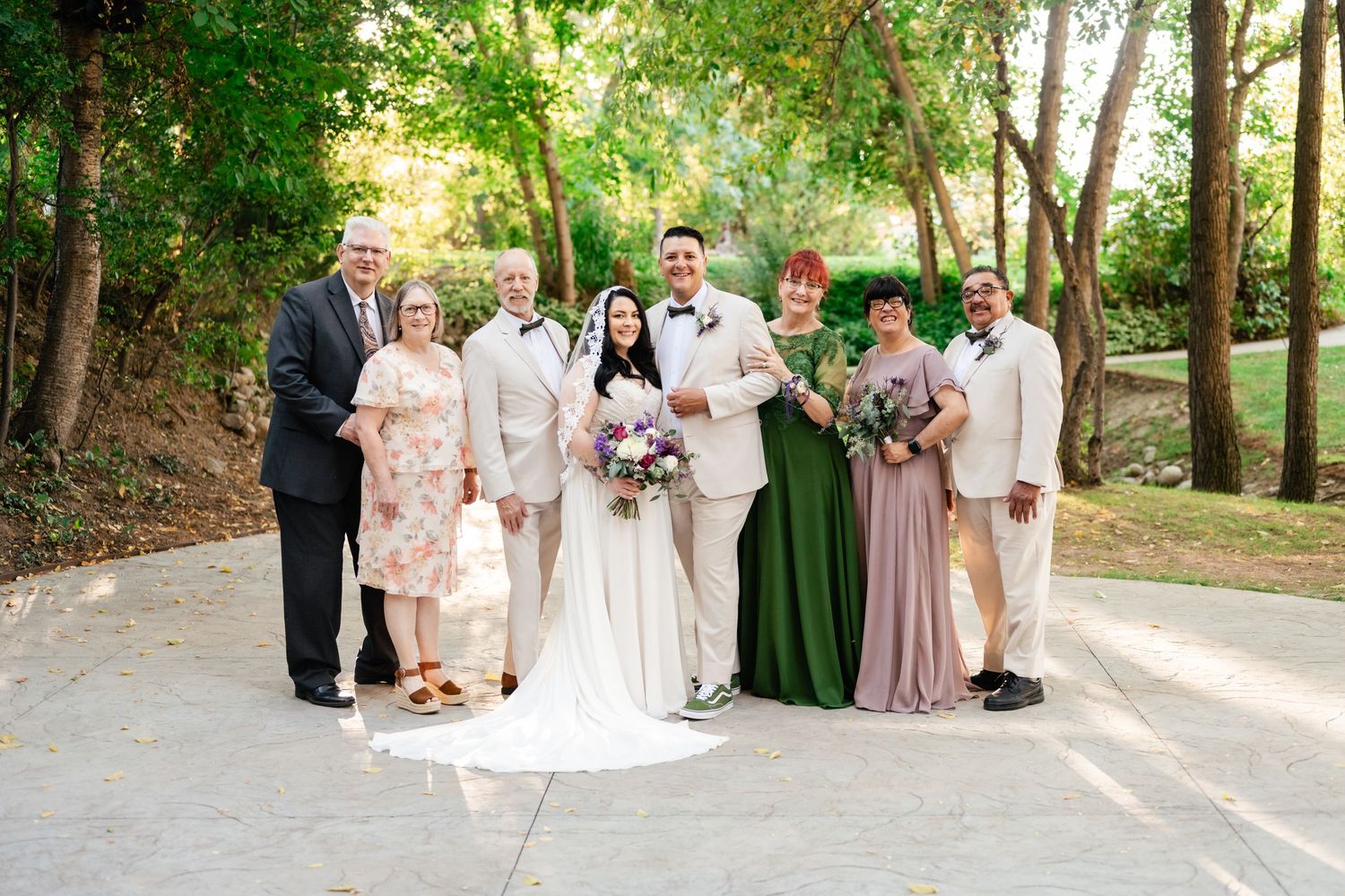 A wedding party group photo with bridesmaids in pastel dresses and groomsmen in light-colored suits outdoors.