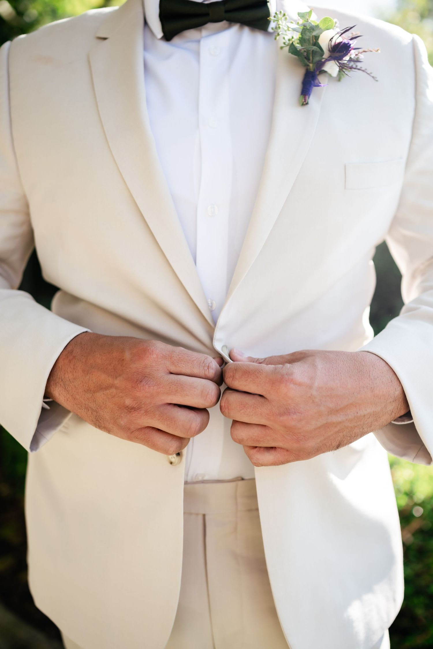 Close-up of groom in white tuxedo jacket and bowtie adjusting buttons with purple boutonniere on lapel.