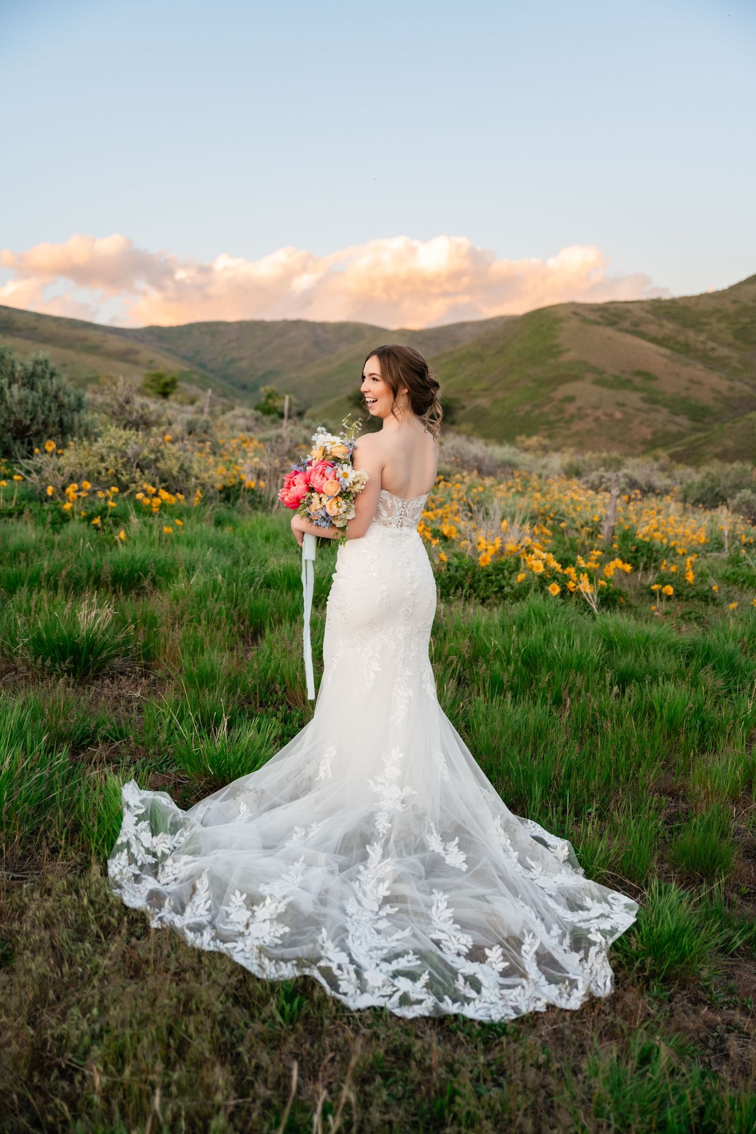 Bride in lace wedding gown holds bouquet while looking over mountain landscape at sunset.