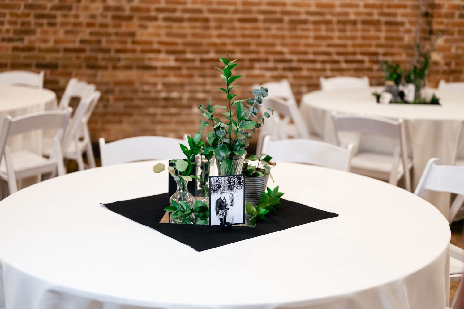 A minimalist wedding centerpiece with green eucalyptus in glass vases on a black napkin atop a white round table.