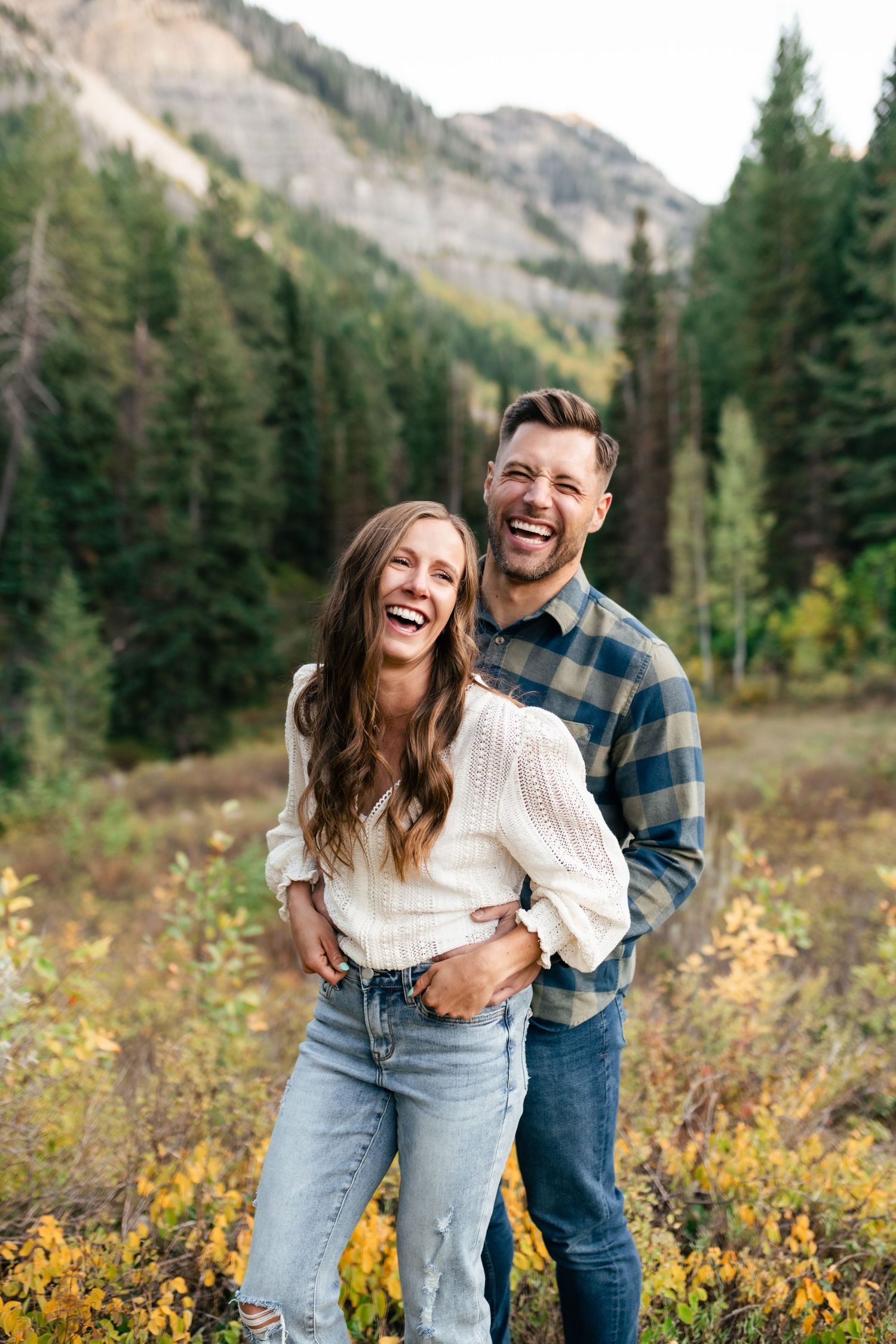Couple laughing together in autumn mountain meadow with evergreen forest and rocky peaks in background.