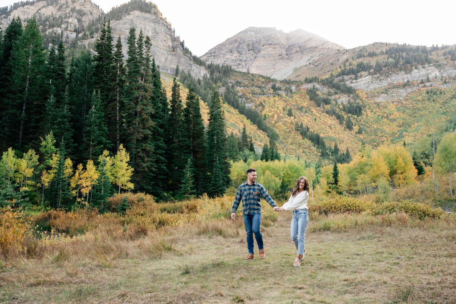 Two people walk together on a mountain trail surrounded by evergreen trees and autumn foliage in Colorado.