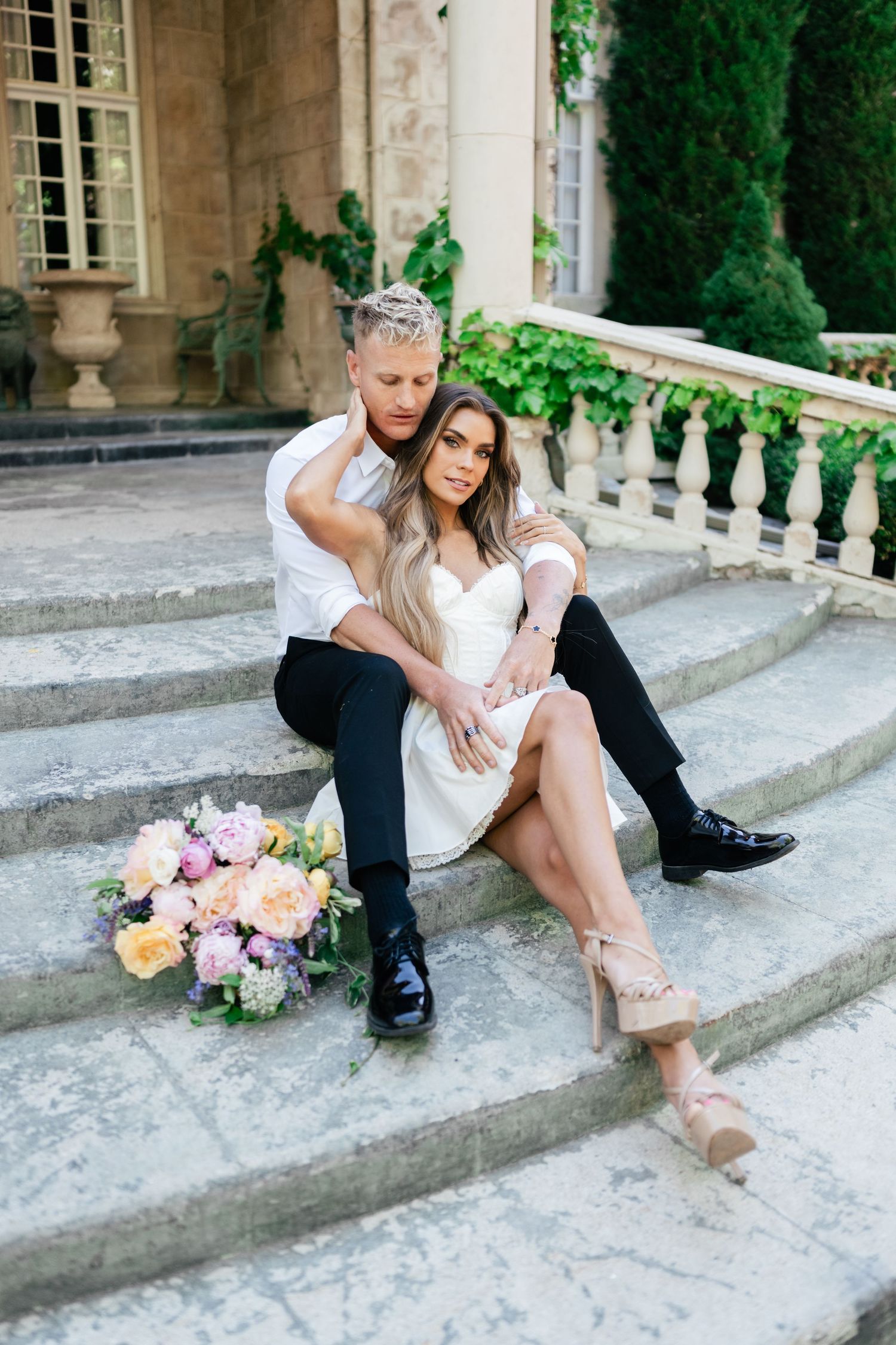 Elegant couple sitting on stone steps outside mansion with colorful floral bouquet in romantic photoshoot setting.