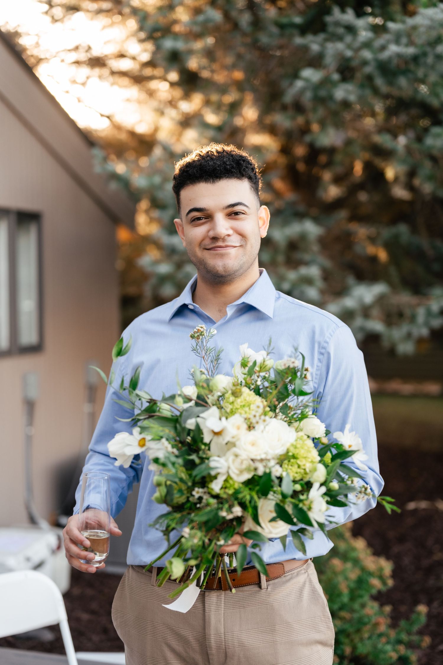 A person in a light blue button-down shirt and khaki pants holds a beautiful white and green floral bouquet outdoors.