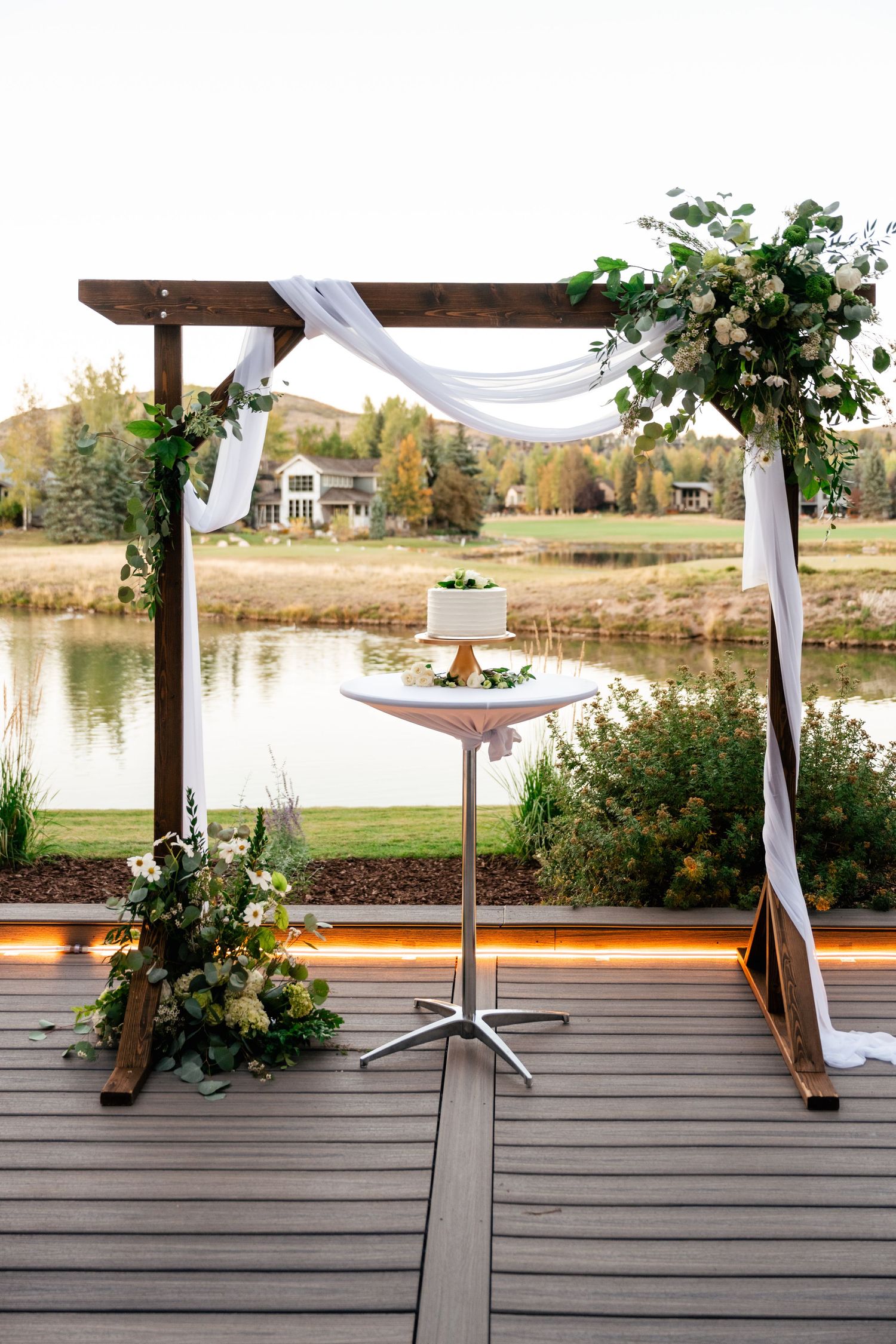 A wooden deck wedding setup with white draping and floral arrangements overlooking a scenic lake view.